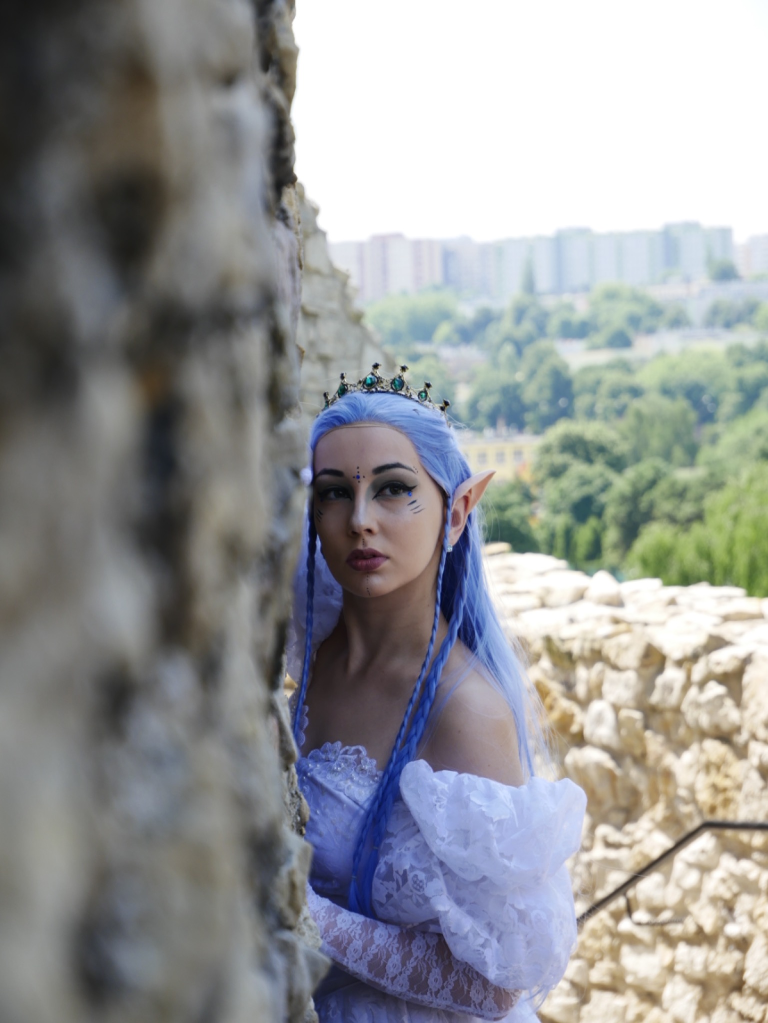 Portret kobiety przebranej za postać z baśni, z niebieskimi włosami, spiczastymi uszami i koroną, pozującej na tle kamiennego muru z widokiem na miasto.