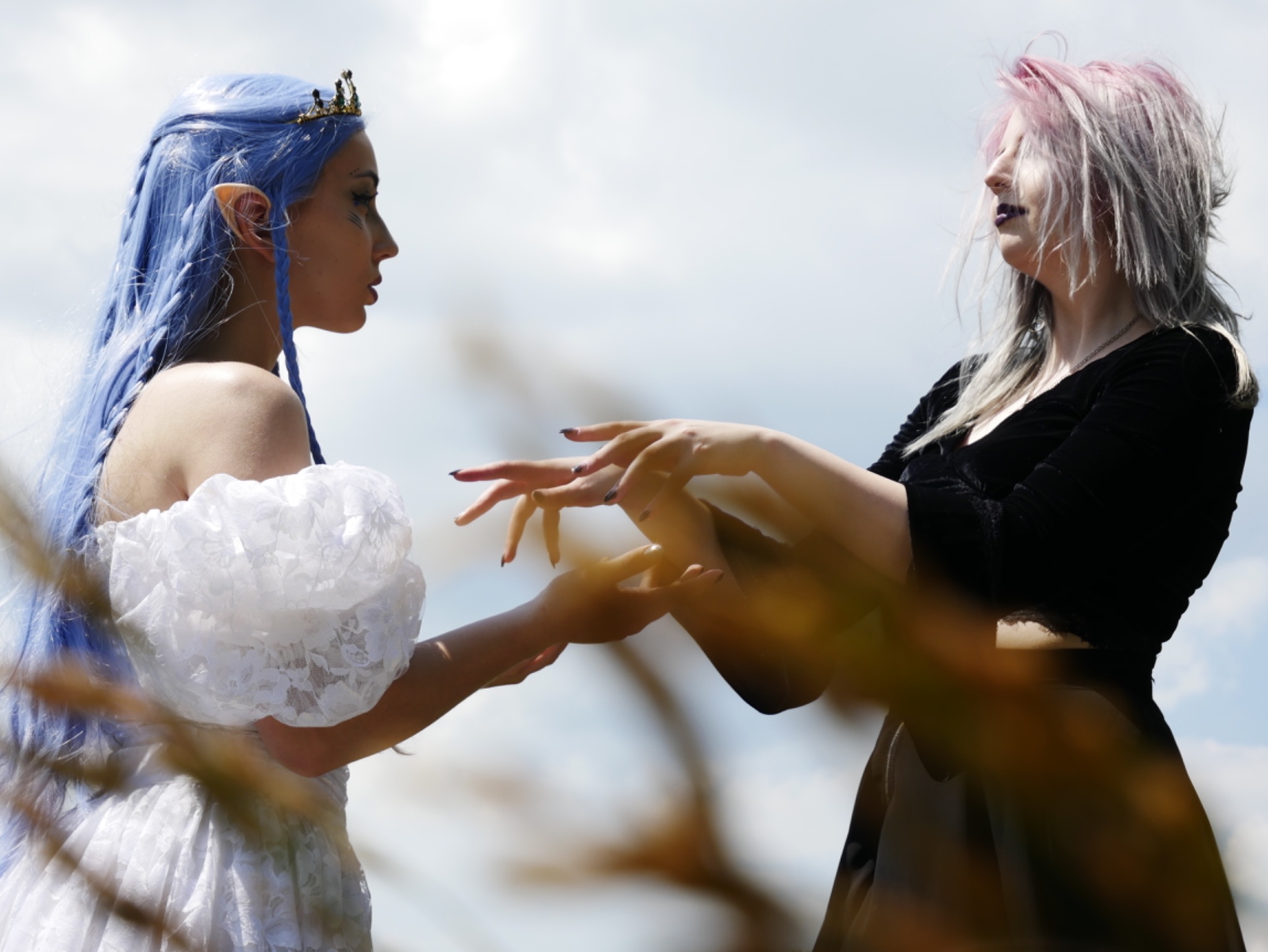 Dwie kobiety w stylizowanych kostiumach i makijażu: jedna z błękitnymi włosami i elfimi uszami w białej sukni, druga z siwymi włosami i ciemnymi ustami w czarnej sukni, zbliżają do siebie dłonie...