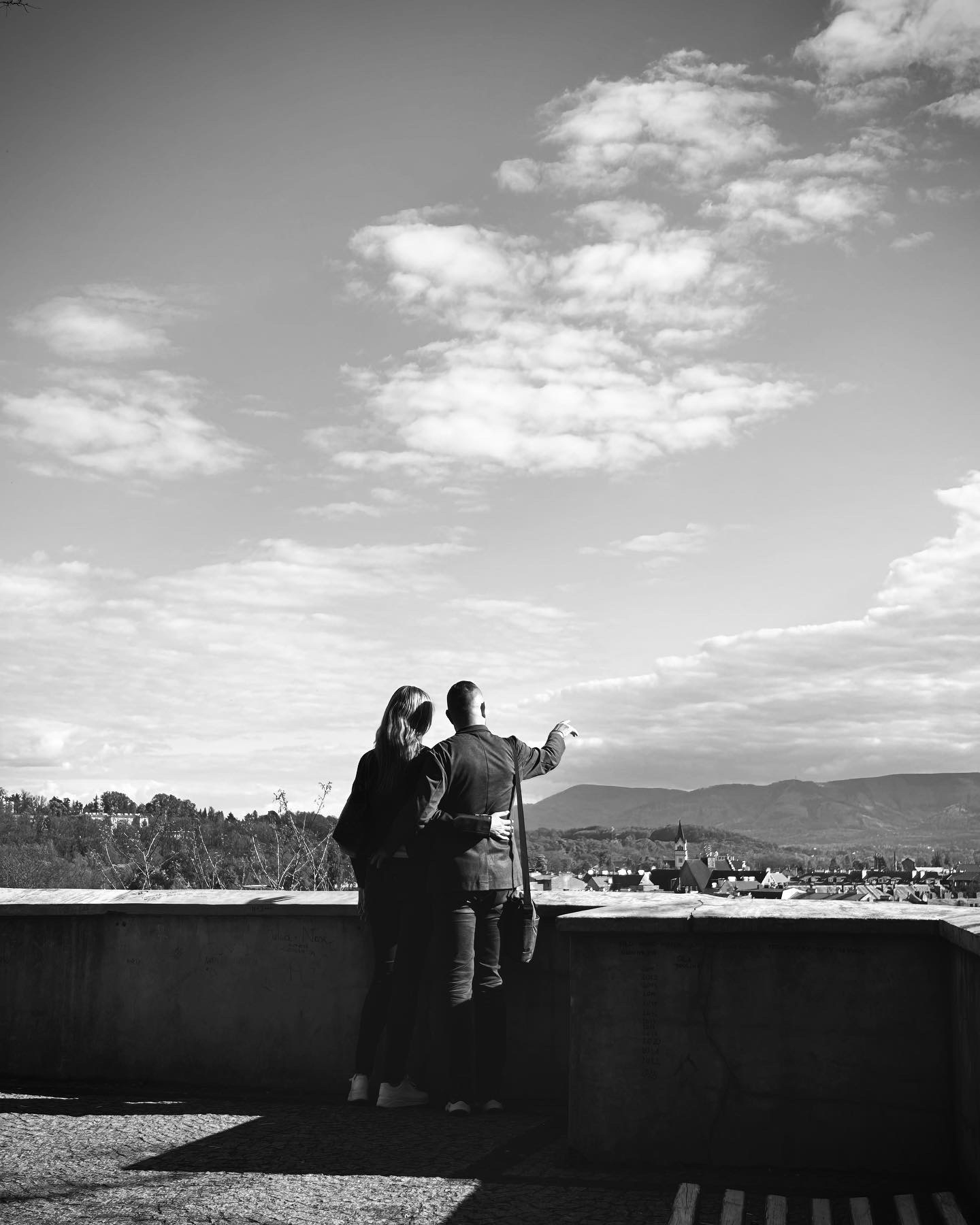 Czarno-białe zdjęcie pary stojącej na tarasie z widokiem na miasto i góry w tle, mężczyzna wskazuje palcem w dal.