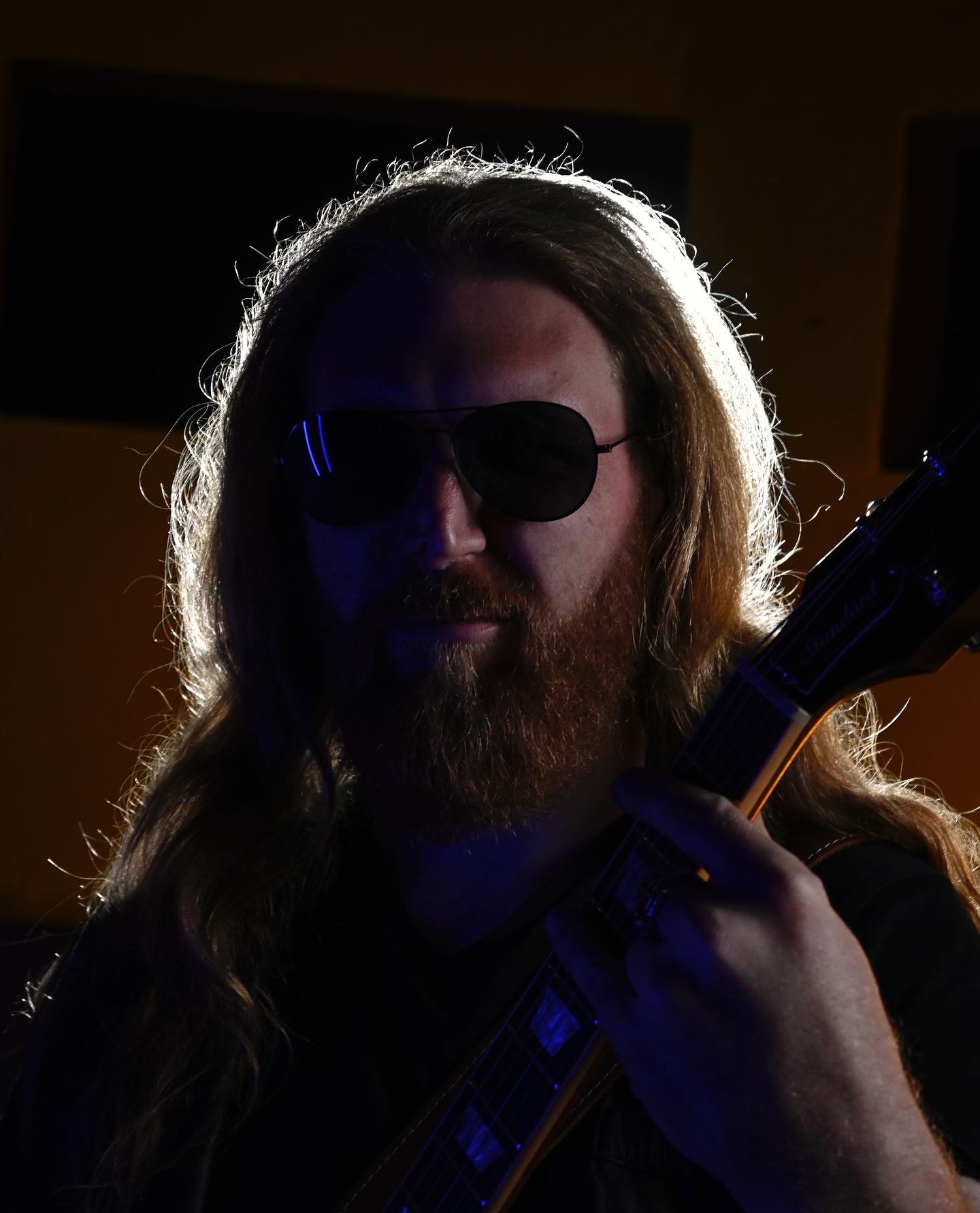 Portret mężczyzny z długimi włosami i brodą w okularach przeciwsłonecznych, trzymającego gitarę elektryczną, oświetlonego od tyłu mocnym światłem kontrującym.