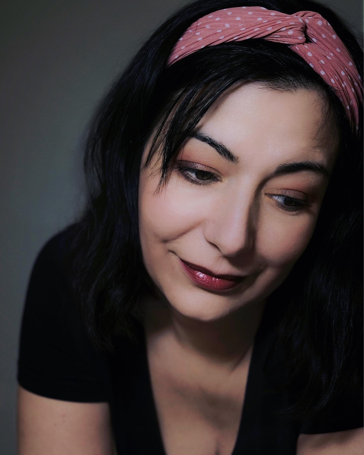 Portret kobiety z ciemnymi włosami i różową opaską w kropki, spoglądającej w dół, z delikatnym makijażem oczu i ust w odcieniach bordo.