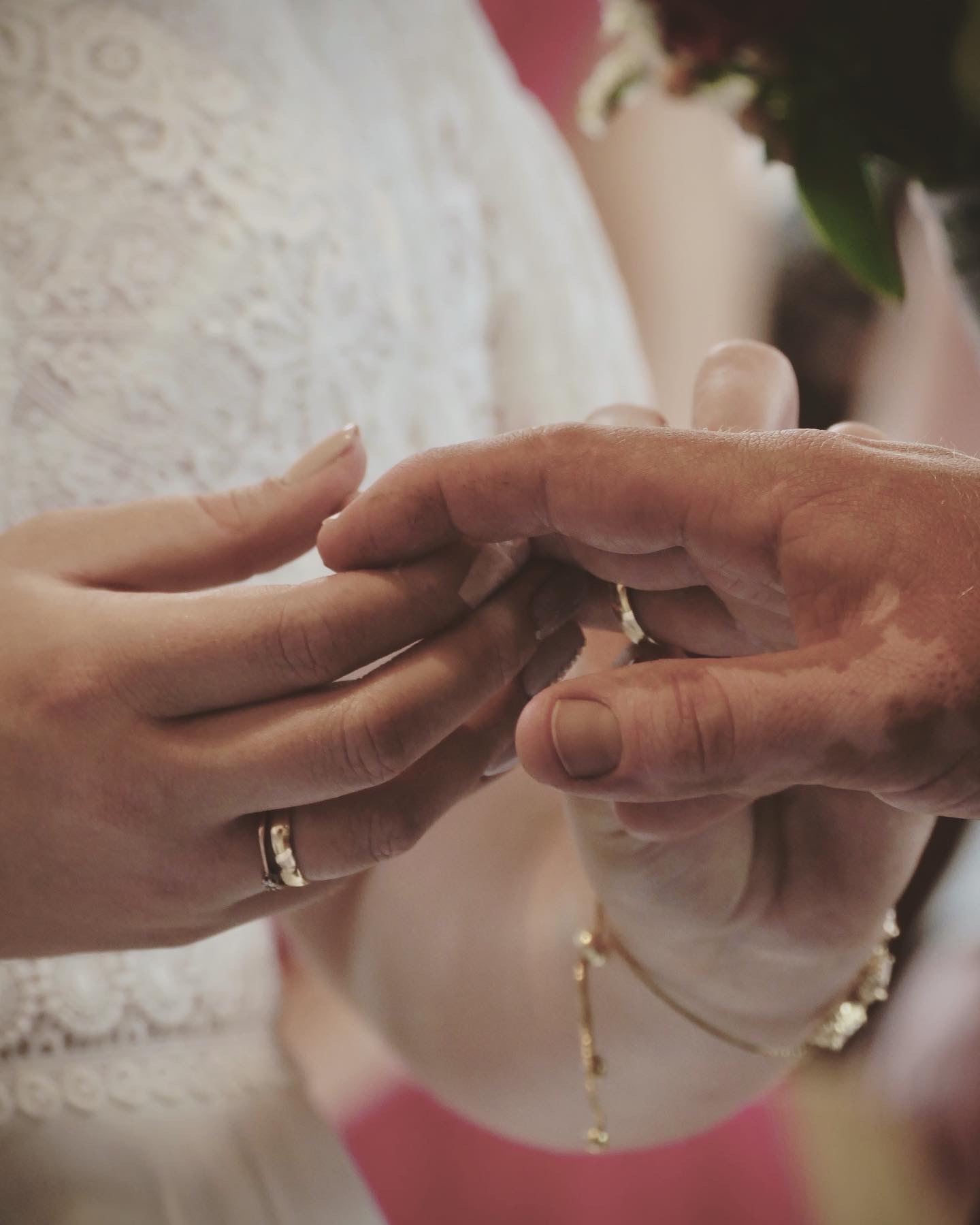 Zbliżenie dłoni w trakcie ceremonii zaślubin, panna młoda zakłada obrączkę na palec pana młodego, widoczna koronkowa suknia i złota bransoletka.