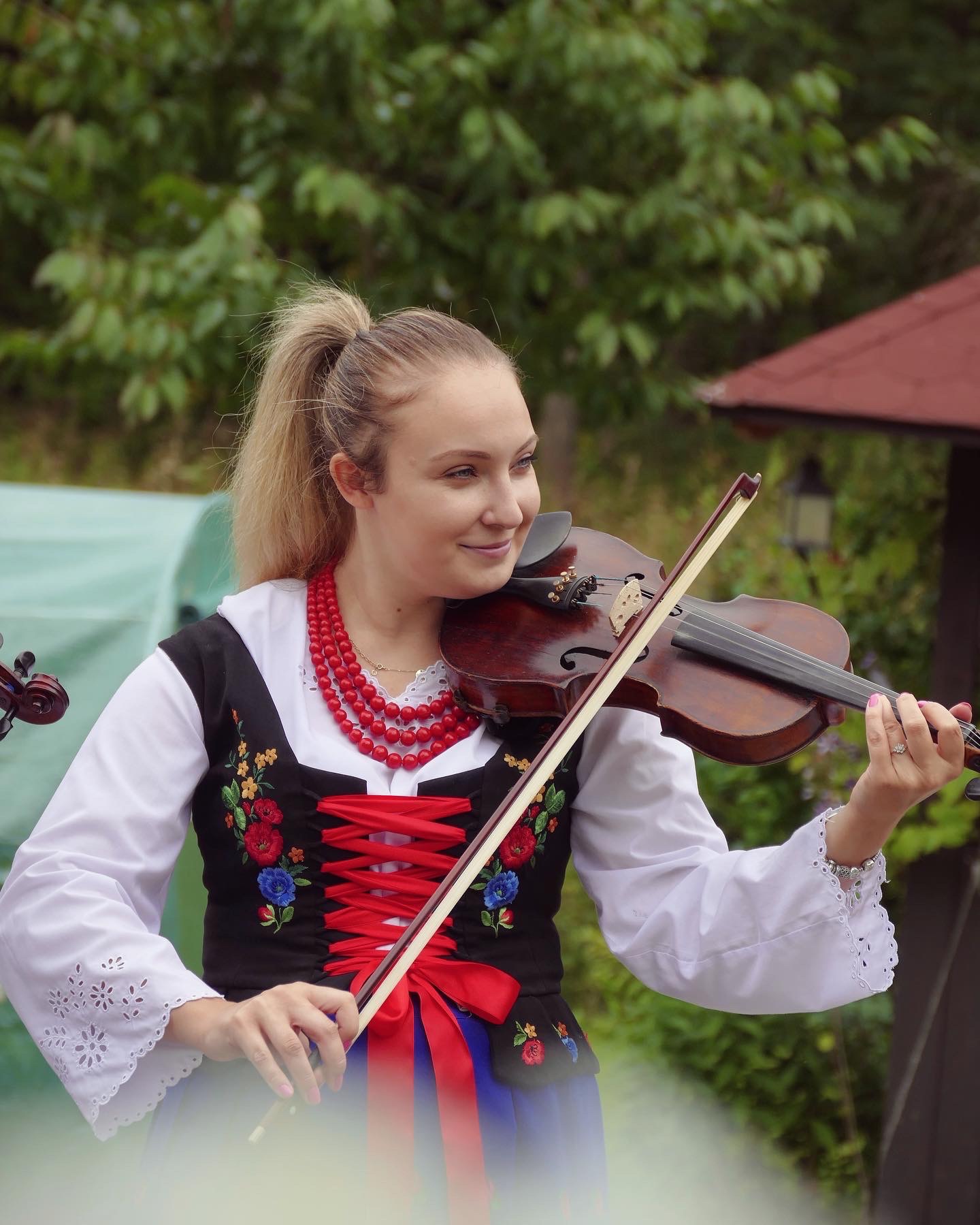 Młoda blondynka w stroju ludowym gra na skrzypcach na tle zieleni.