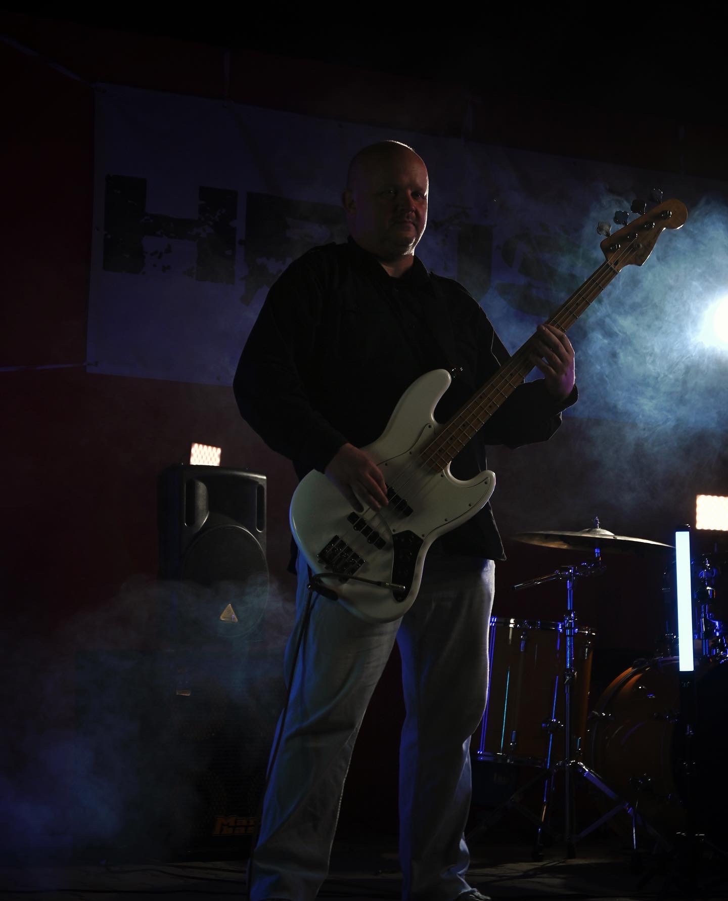 Muzyk grający na białej gitarze basowej na scenie z perkusją i efektami świetlnymi.