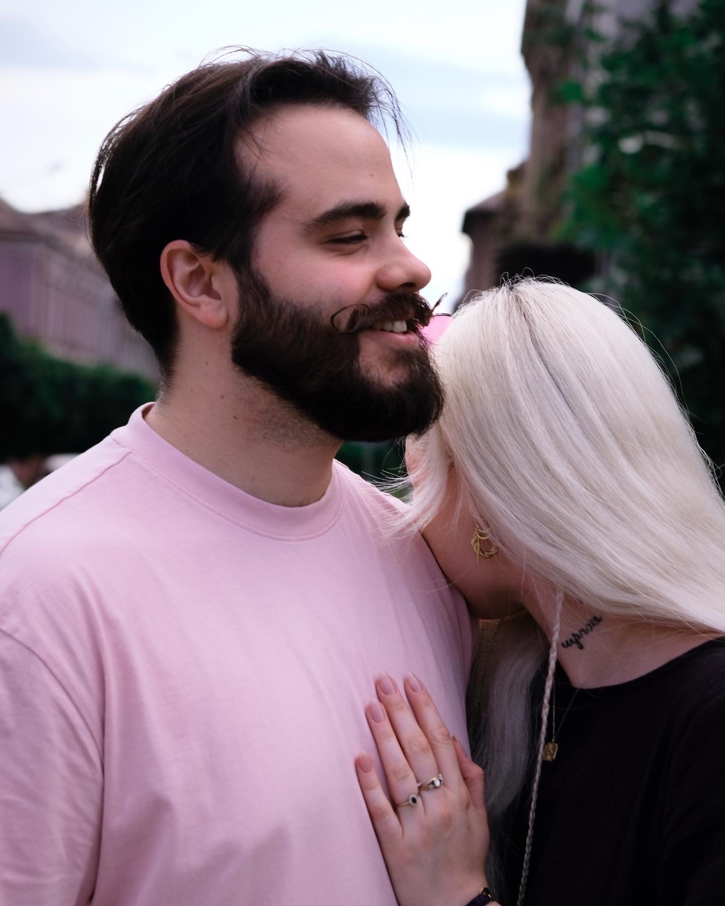 Uśmiechnięty mężczyzna z brodą i wąsami w różowej koszulce, obejmowany przez kobietę z platynowymi włosami i tatuażem na szyi, widoczne złote kolczyki i pierścionki.