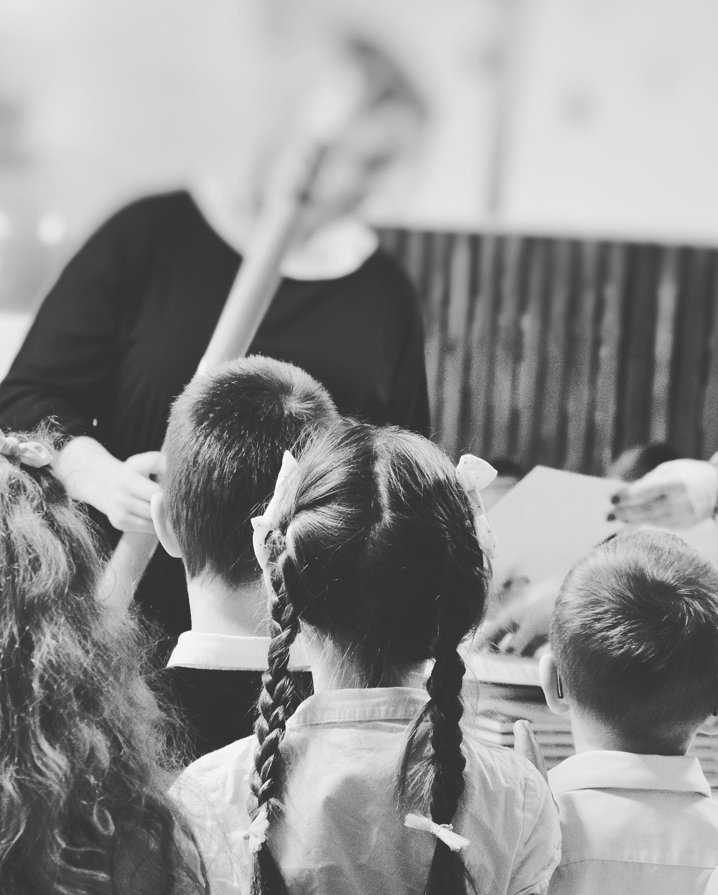 Czarno-białe zdjęcie z tyłu przedstawiające dzieci podczas sesji zdjęciowej, gdzie fotografka robi im zdjęcia, a dziewczynka ma zaplecione warkocze z kokardami.