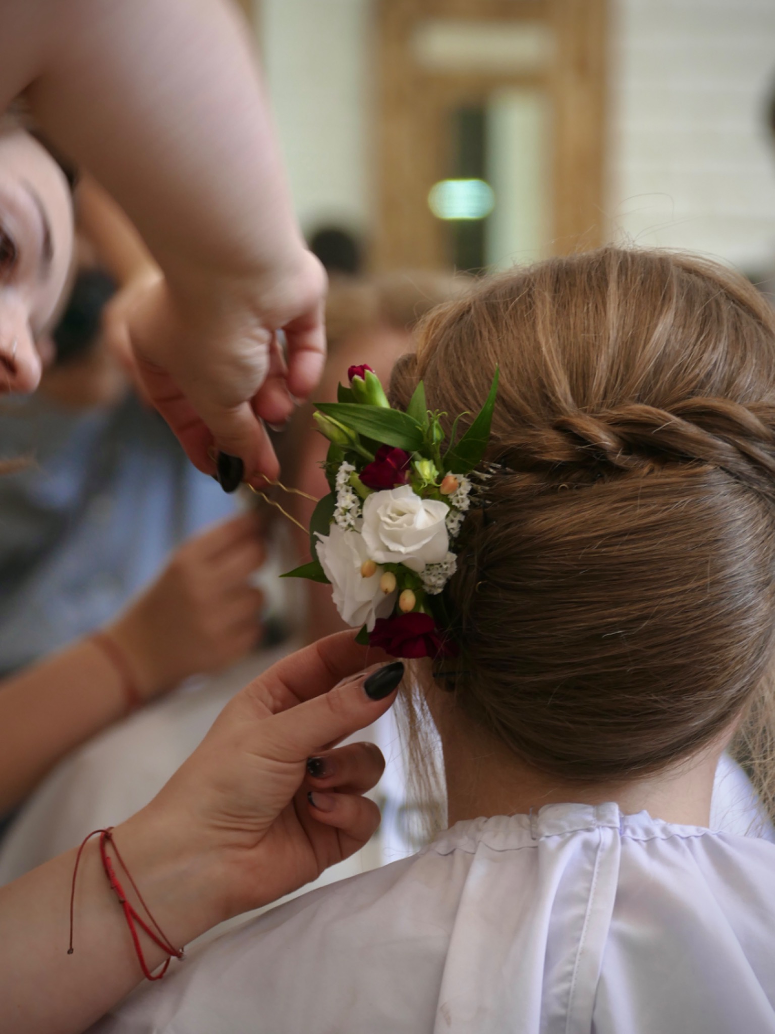 Fryzura ślubna z wplecionymi białymi i czerwonymi kwiatami, upinana przez stylistkę, widok z tyłu, delikatny warkocz.