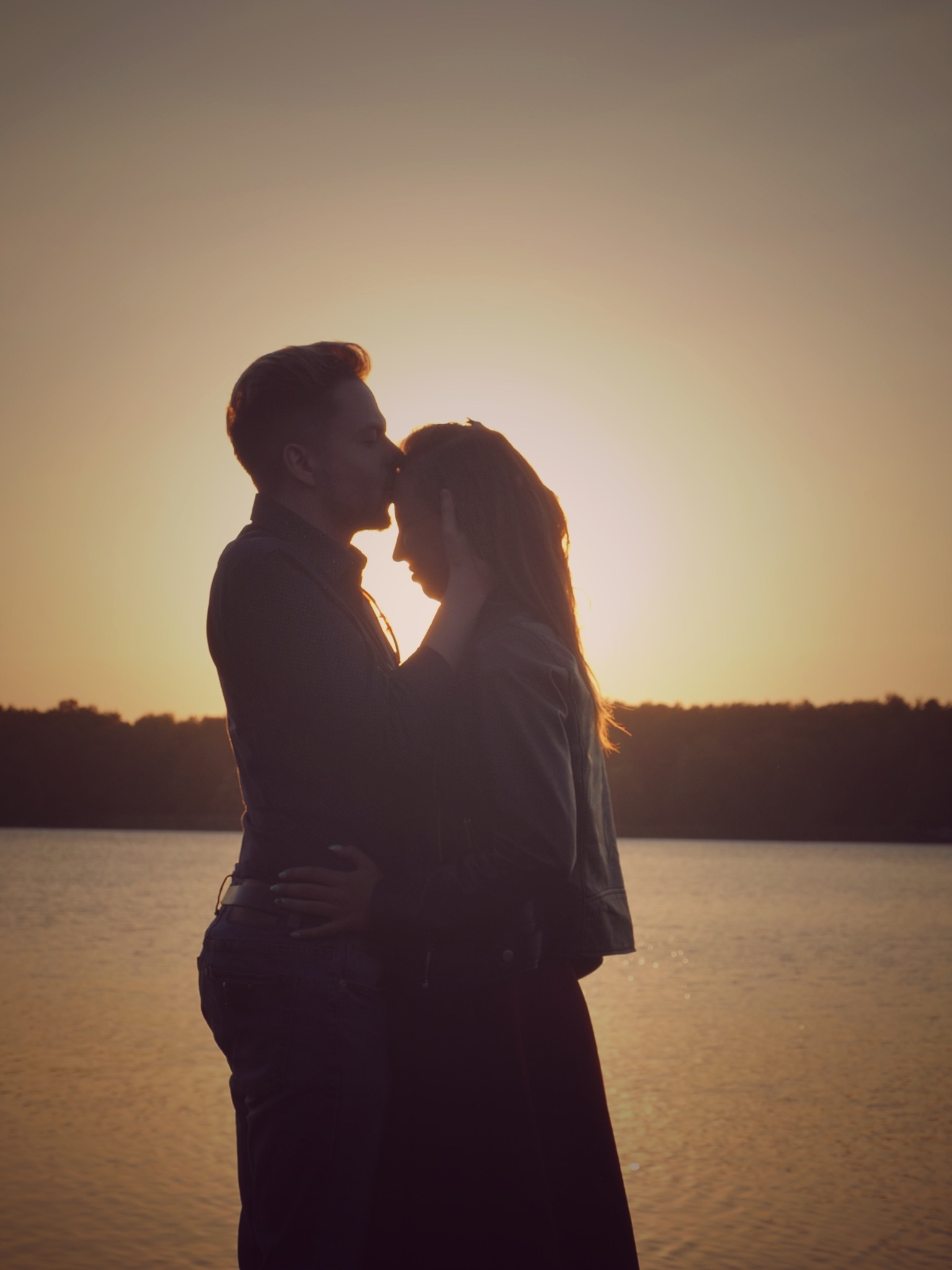 Silhouettes of a couple embracing against a golden sunset over a calm lake; the man kisses the woman's forehead, creating a romantic and intimate moment.