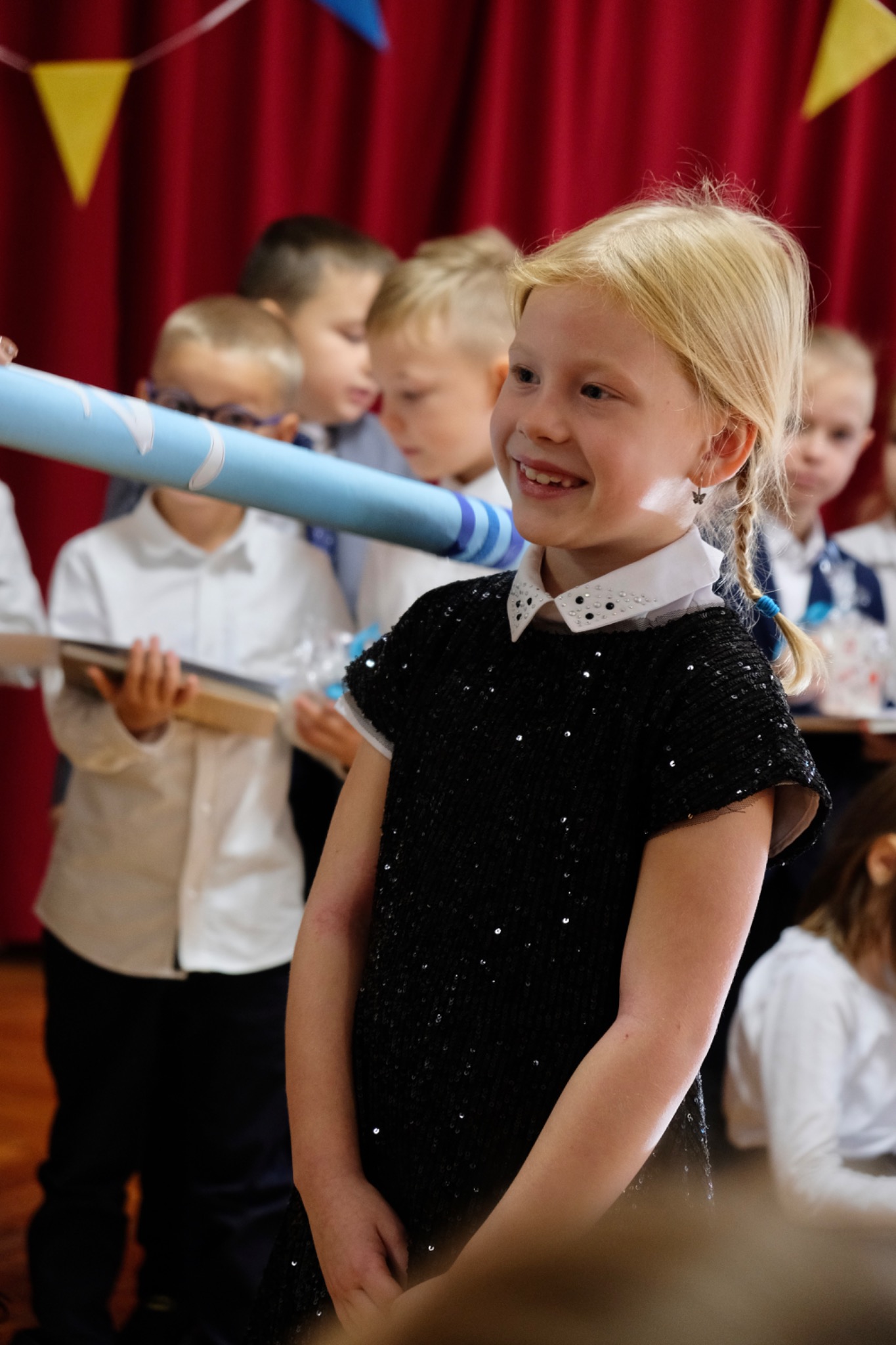 Uśmiechnięta blond dziewczynka w czarnej, błyszczącej sukience z białym kołnierzykiem na tle grupy dzieci i czerwonej zasłony.