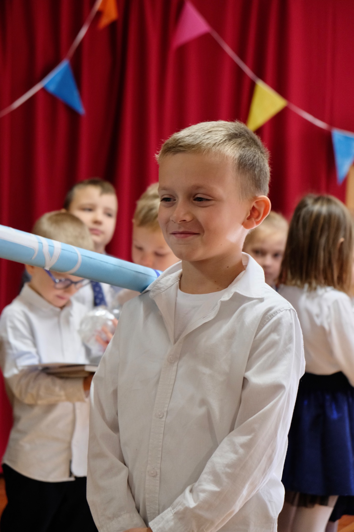 Chłopiec w białej koszuli, uśmiechnięty, trzyma dyplom w niebieskiej tubie na tle czerwonej kotary i innych dzieci podczas uroczystości.