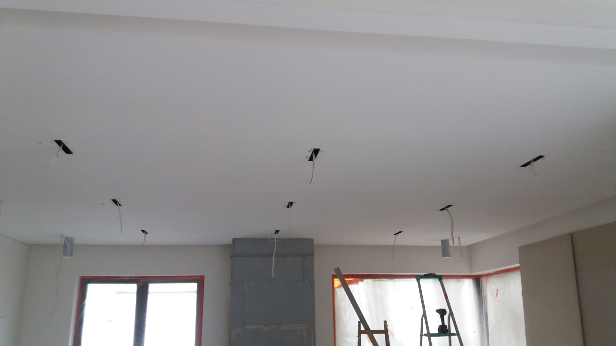 Biały sufit z otworami na oświetlenie, widoczne kable, przygotowany do montażu lamp, w tle okno zabezpieczone folią i drabina.