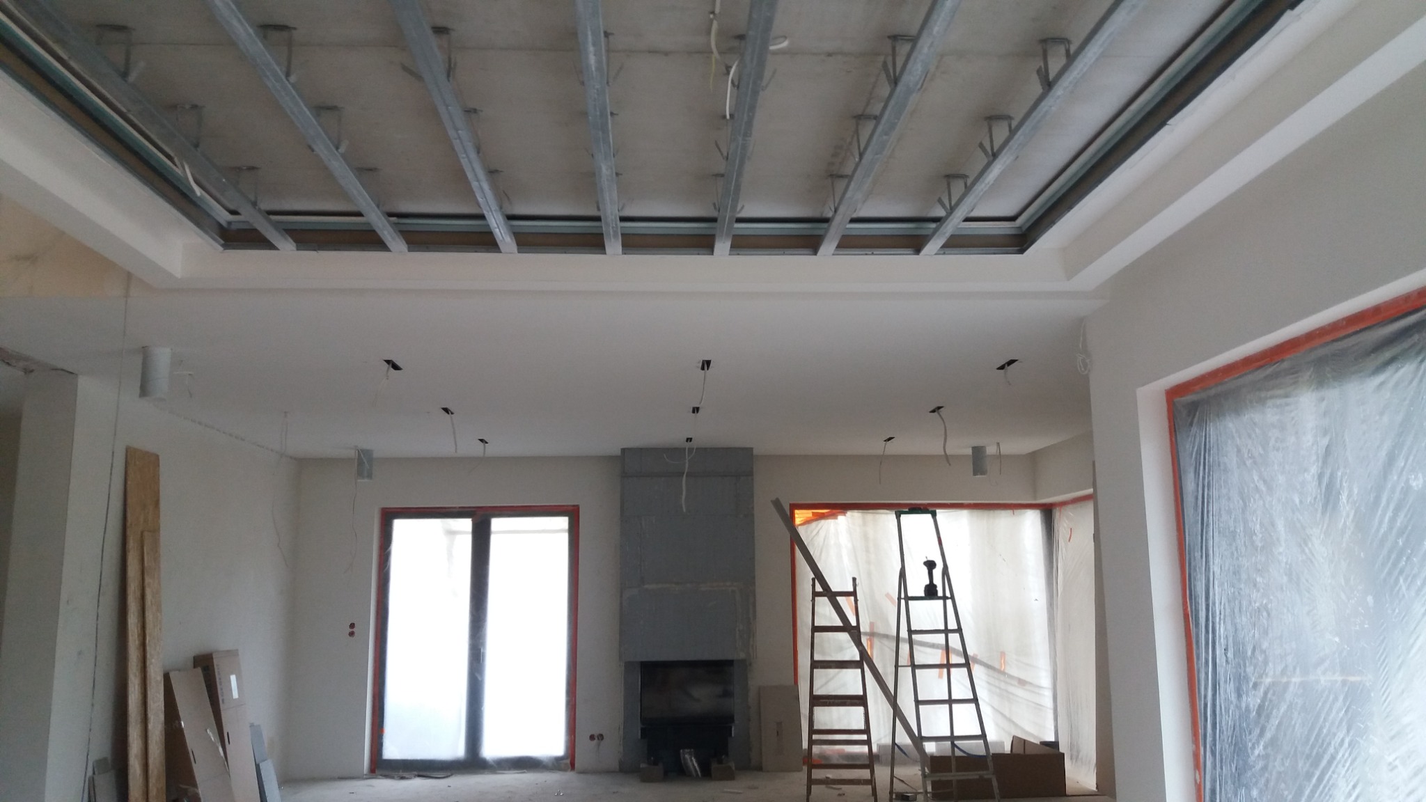 Wnętrze w trakcie prac wykończeniowych: widoczna konstrukcja sufitu podwieszanego z metalowych profili, przygotowana do montażu płyt, oraz ściany po wstępnym wyrównaniu, zabezpieczone okna folią...