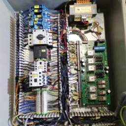 Wnętrze szafy sterowniczej z widocznymi elementami elektrycznymi, w tym przekaźnikami, bezpiecznikami, listwami zaciskowymi i przewodami o różnych kolorach, uporządkowanymi wewnątrz metalowej obudowy.