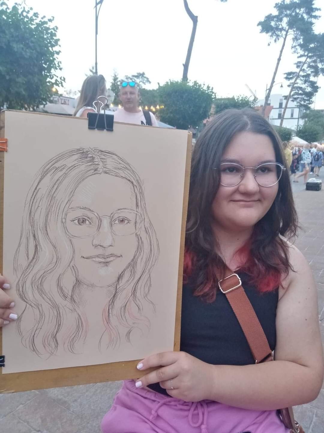 Portret ołówkiem kobiety w okularach, trzymany przez modelkę z czerwonymi pasemkami na włosach, na tle miejskiego placu.