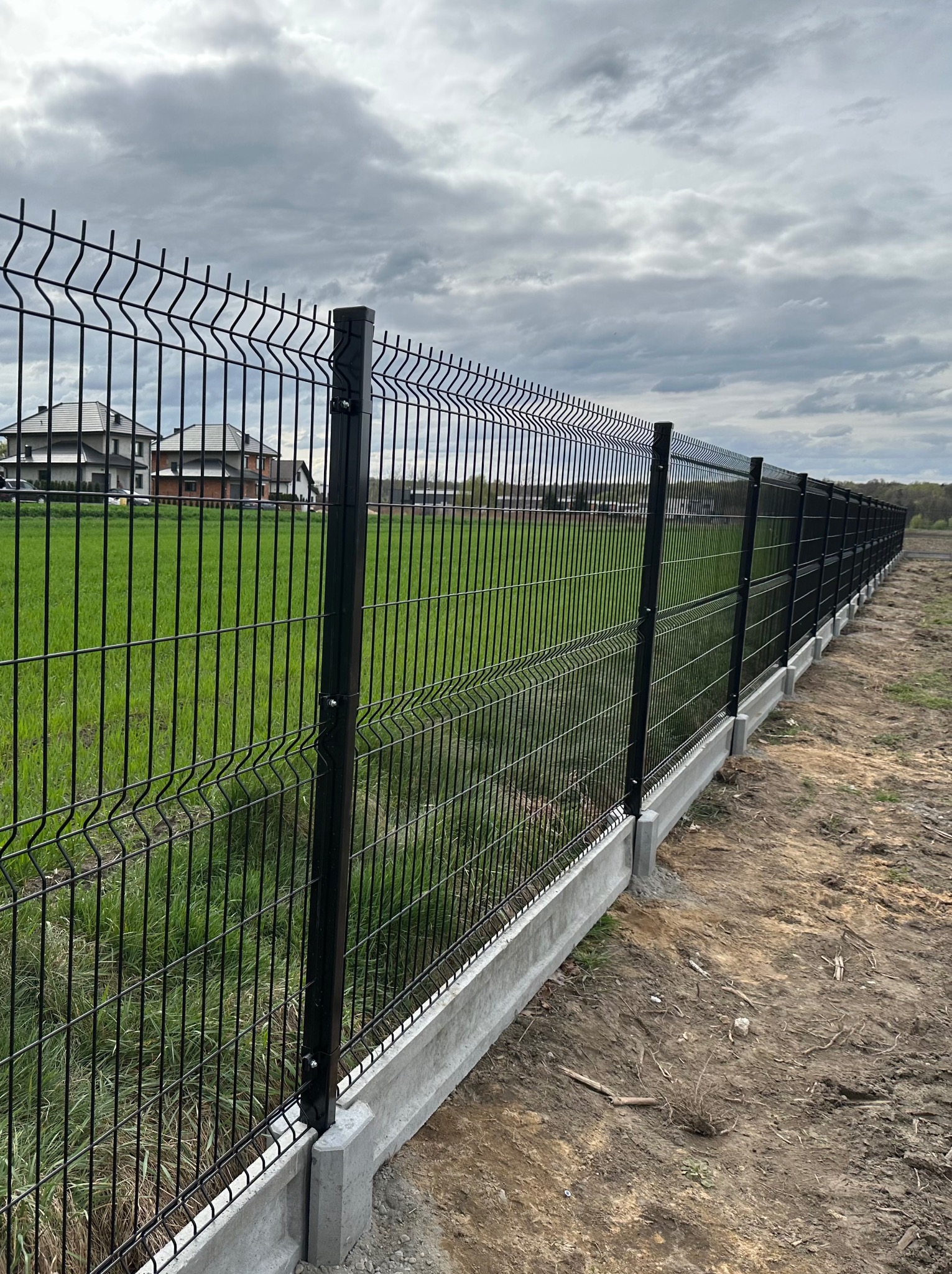 Długi, czarny, metalowy płot panelowy z betonową podmurówką na nierównym terenie, w tle trawnik i domy pod pochmurnym niebem.