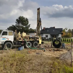 Wiertnica na podwoziu ciężarówki podczas pracy na działce, widoczny wykop, rury, budynek mieszkalny w tle, pochmurne niebo.