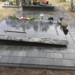 Nowo ułożona, ciemna kostka brukowa otaczająca nagrobek z szarego kamienia z napisem w złotych literach, dekoracje kwiatowe i lampiony.