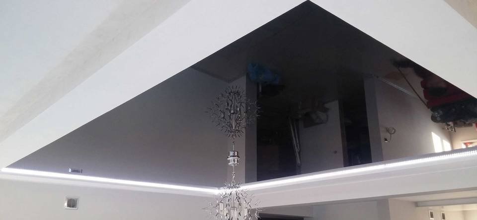 Błyszczący, czarny sufit napinany w trójkątnym kształcie, z oświetleniem LED i odbijającym się wnętrzem pokoju, zwieńczony ozdobnym żyrandolem.