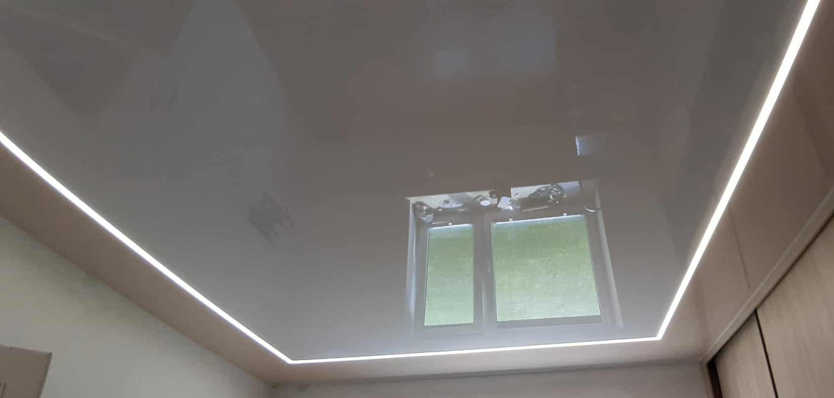 Błyszczący, szary sufit napinany z oświetleniem LED wzdłuż krawędzi, odbijający okno. Realizacja w Tarnowie. Nowoczesne wykończenie wnętrz.