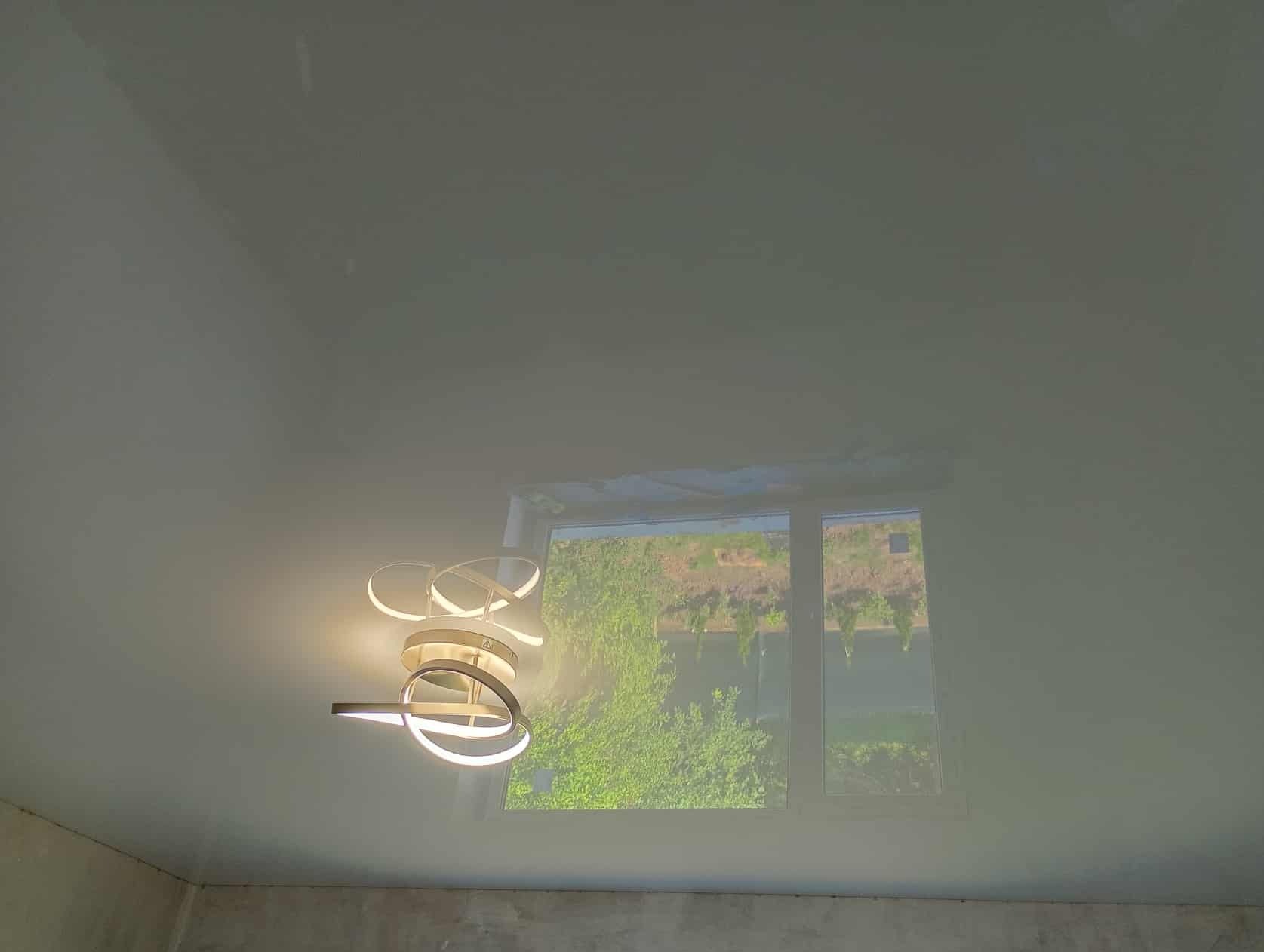 Biały sufit napinany z nowoczesną lampą LED o spiralnym kształcie, widok z dołu, okno z zielenią w tle. Minimalistyczny design wnętrza.