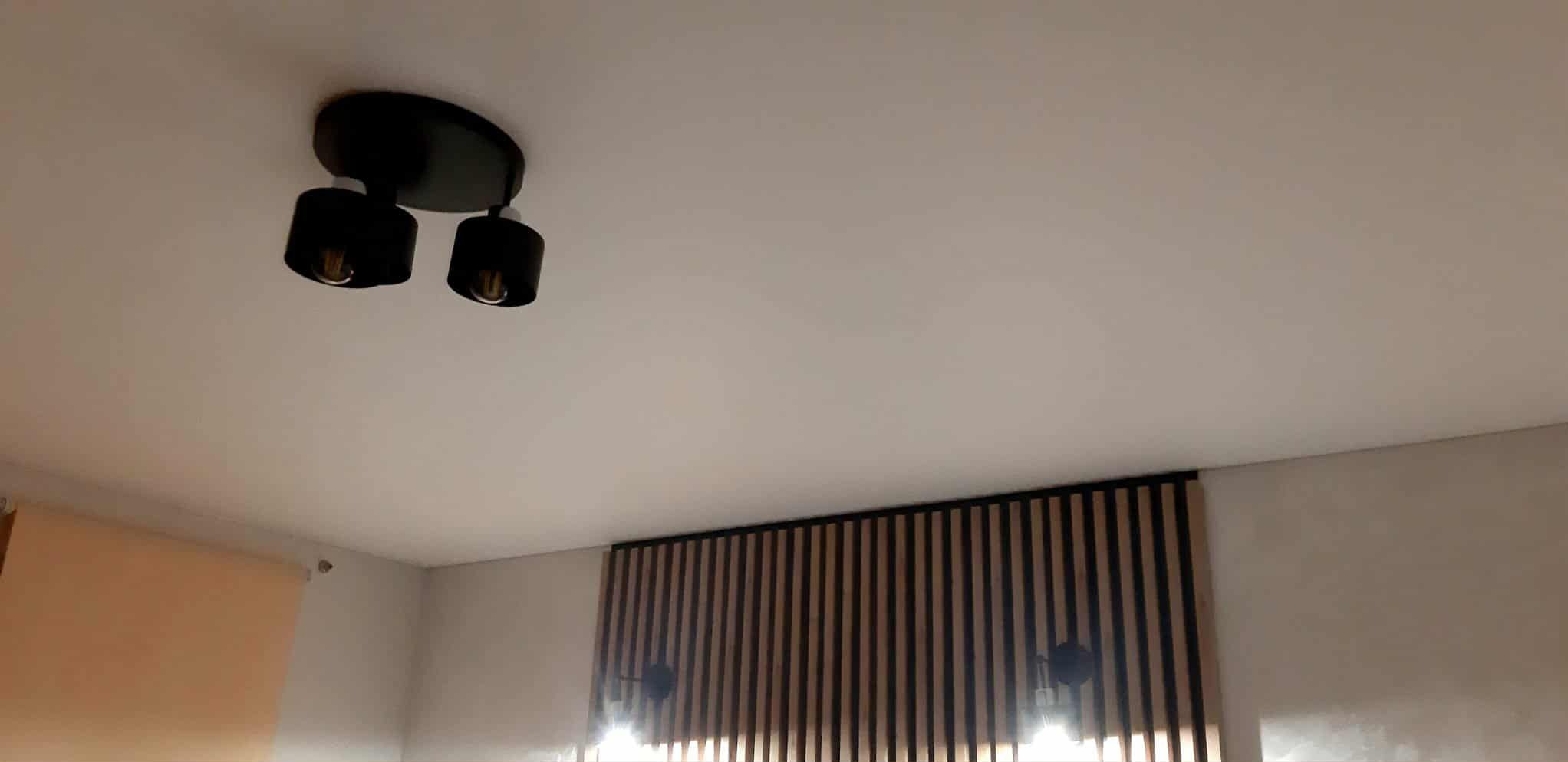 Biały sufit z czarną, nowoczesną lampą dwupunktową. Dekoracyjna ściana z drewnianych listewek i kinkietami. Minimalistyczny design wnętrza.