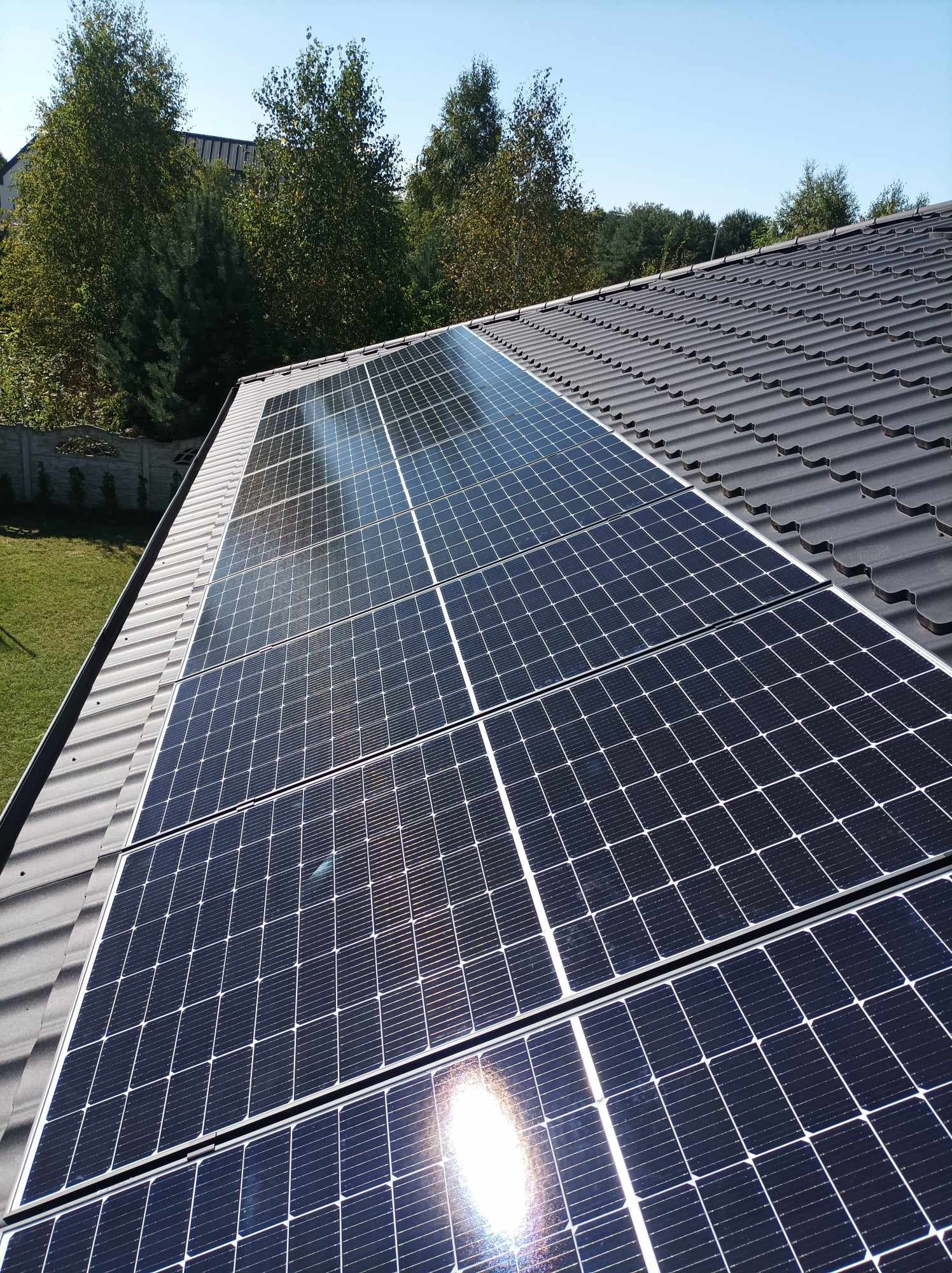 Panel słoneczny na dachu z dachówki ceramicznej w słoneczny dzień, widok z boku na zielony trawnik i drzewa w tle.