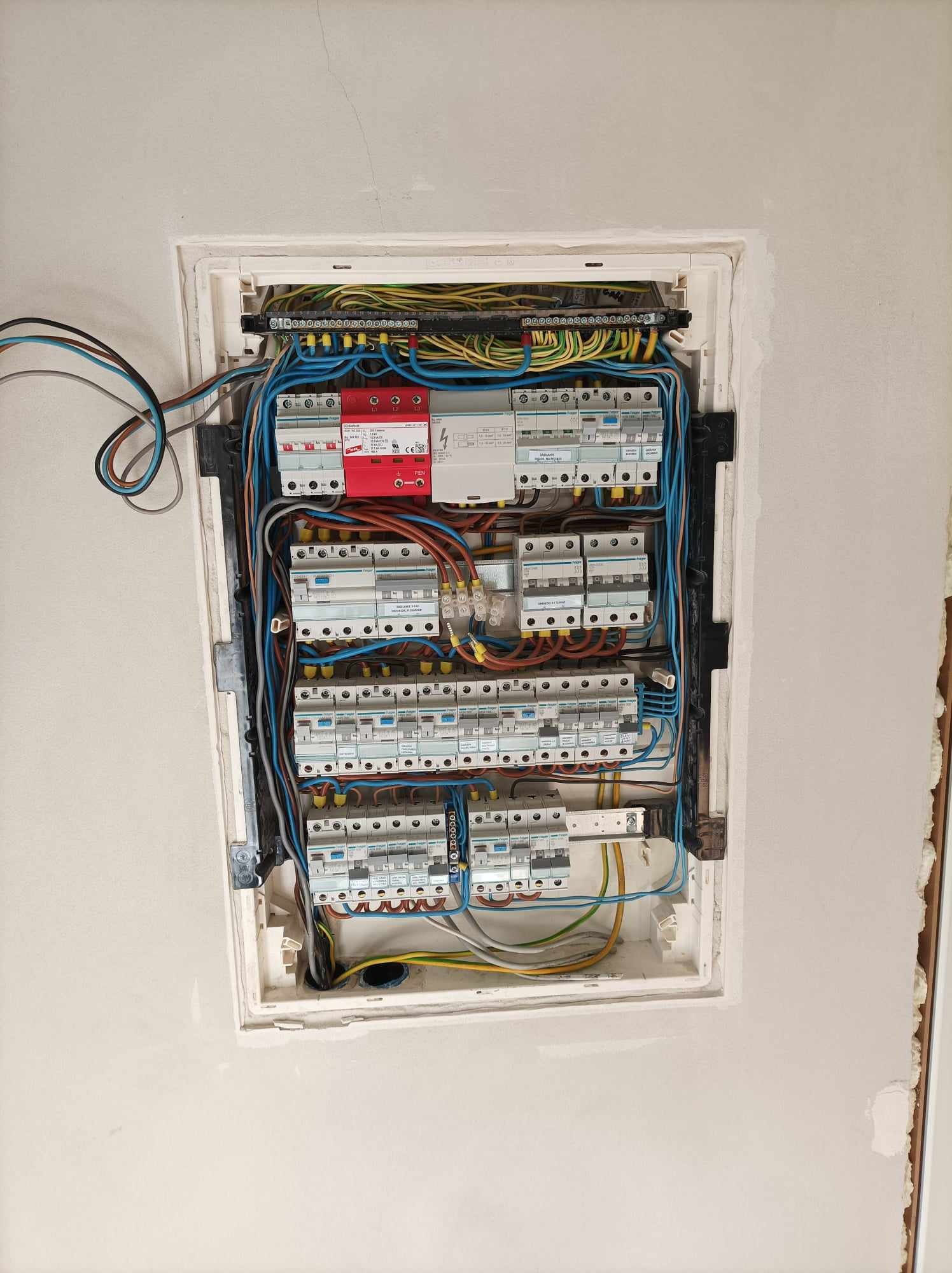 Rozdzielnica elektryczna z zabezpieczeniami różnicowoprądowymi i nadprądowymi, widoczne przewody w różnych kolorach, zamontowana w ścianie z widocznymi śladami szpachlowania.