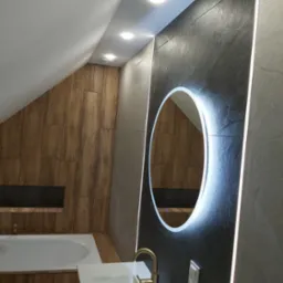 Nowoczesna łazienka na poddaszu z oświetlonym lustrem, drewnianą zabudową wanny i szarą ścianą z płytek. Umywalka z baterią w kolorze złota.