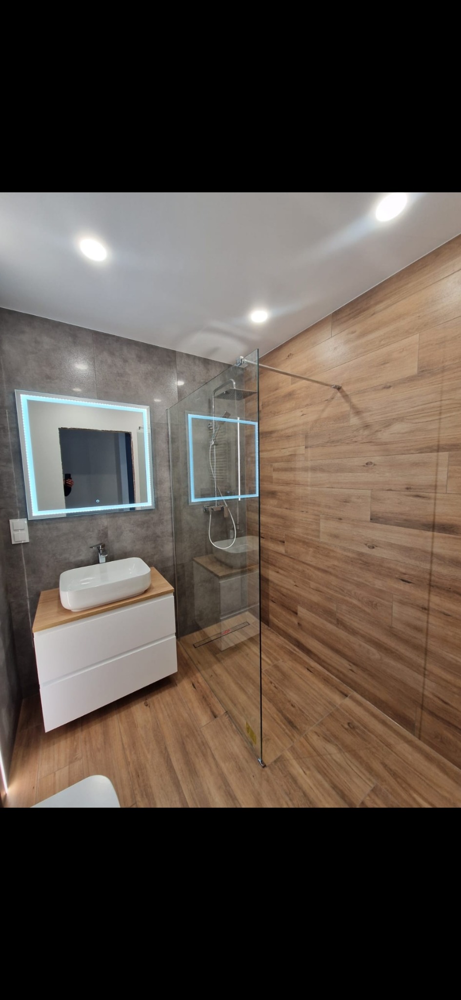 Nowoczesna łazienka z drewnopodobnymi płytkami, kabiną prysznicową typu walk-in ze szklaną ścianką i podświetlanym lustrem nad umywalką nablatową.