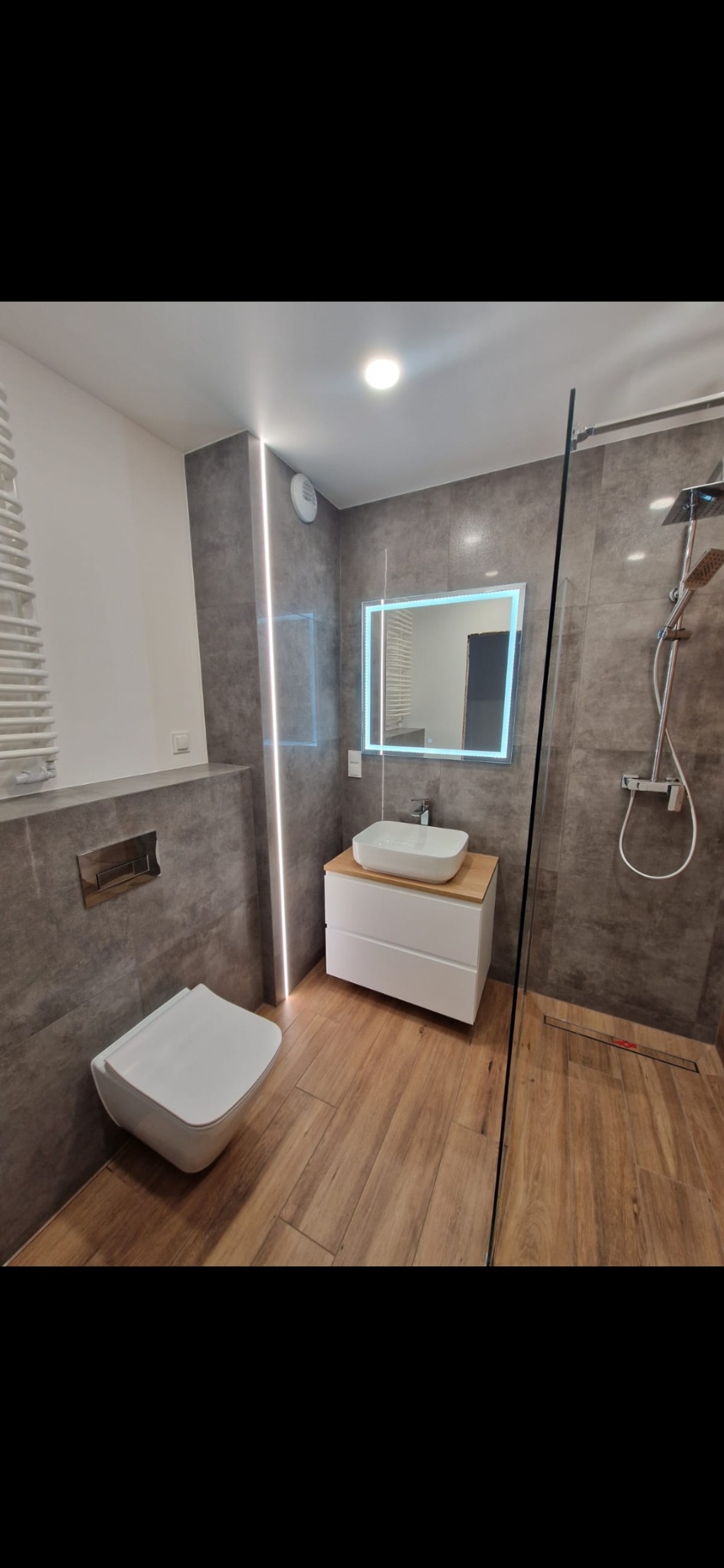 Nowoczesna łazienka z szarymi płytkami, drewnopodobną podłogą, podświetlanym lustrem, wiszącą toaletą i kabiną prysznicową ze szklaną ścianką.