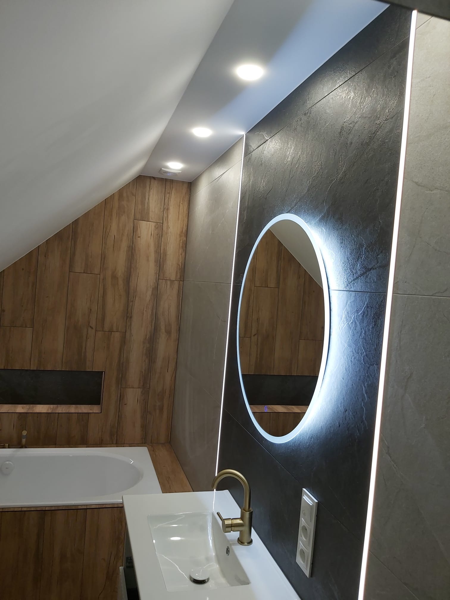 Nowoczesna łazienka na poddaszu z oświetlonym lustrem, drewnianą zabudową wanny i szarą ścianą z płytek. Umywalka z baterią w kolorze złota.