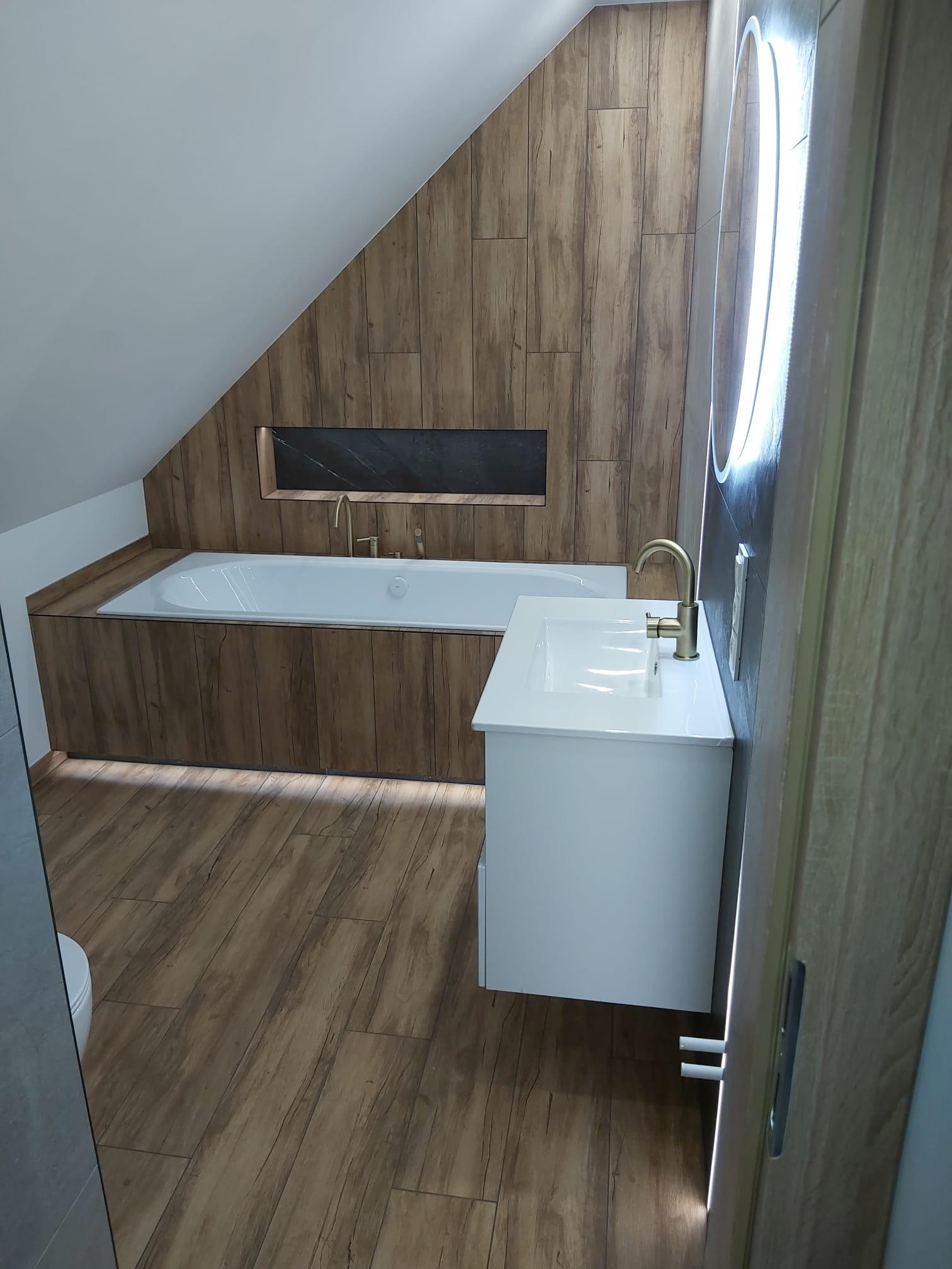 Nowoczesna łazienka na poddaszu z drewnopodobną okładziną, białą wanną i umywalką. Złota armatura i okrągłe lustro z podświetleniem LED.