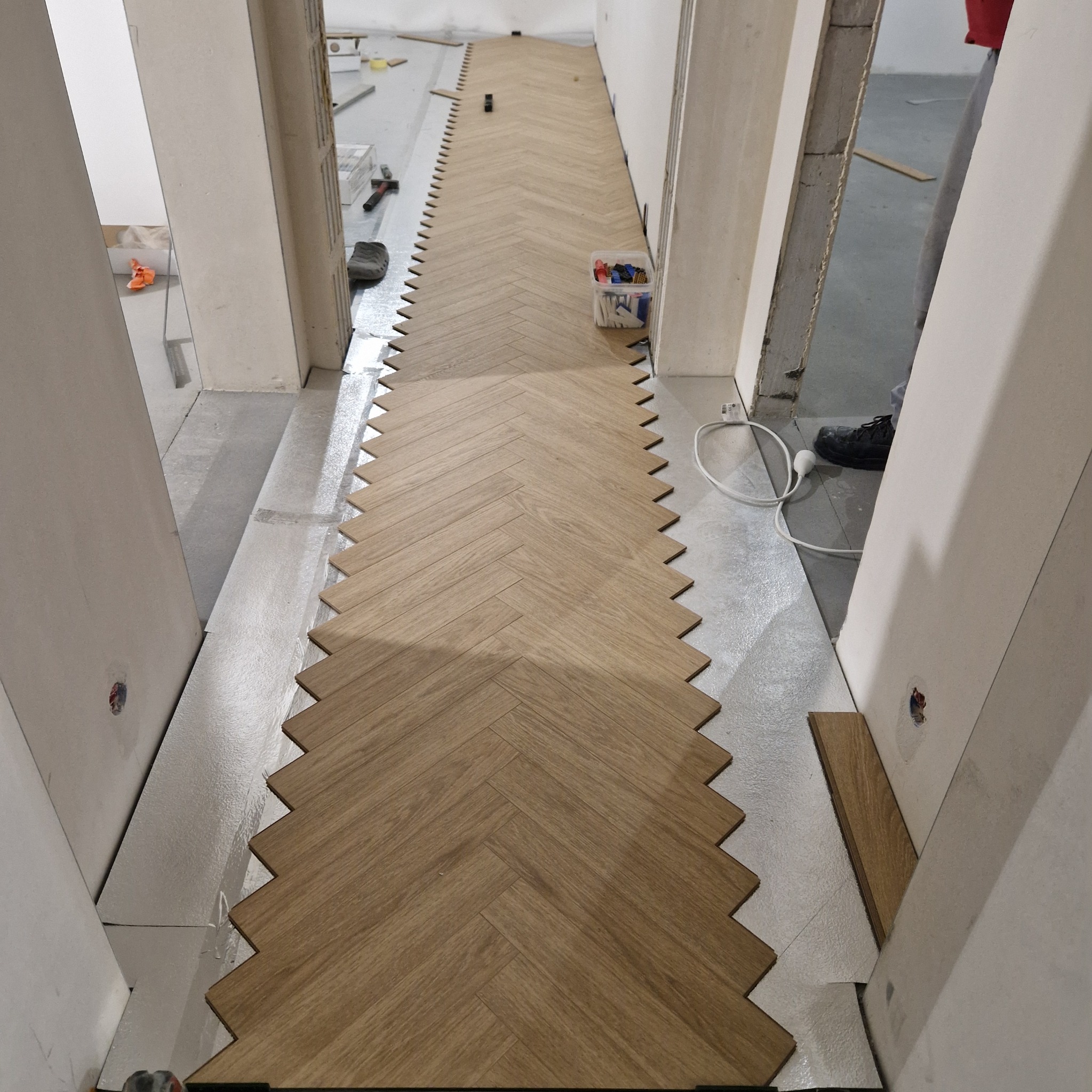 Układanie parkietu w jodełkę w korytarzu w Kętach. Widoczne narzędzia i akcesoria glazurnicze. Parkiet ułożony częściowo na srebrnej folii izolacyjnej.