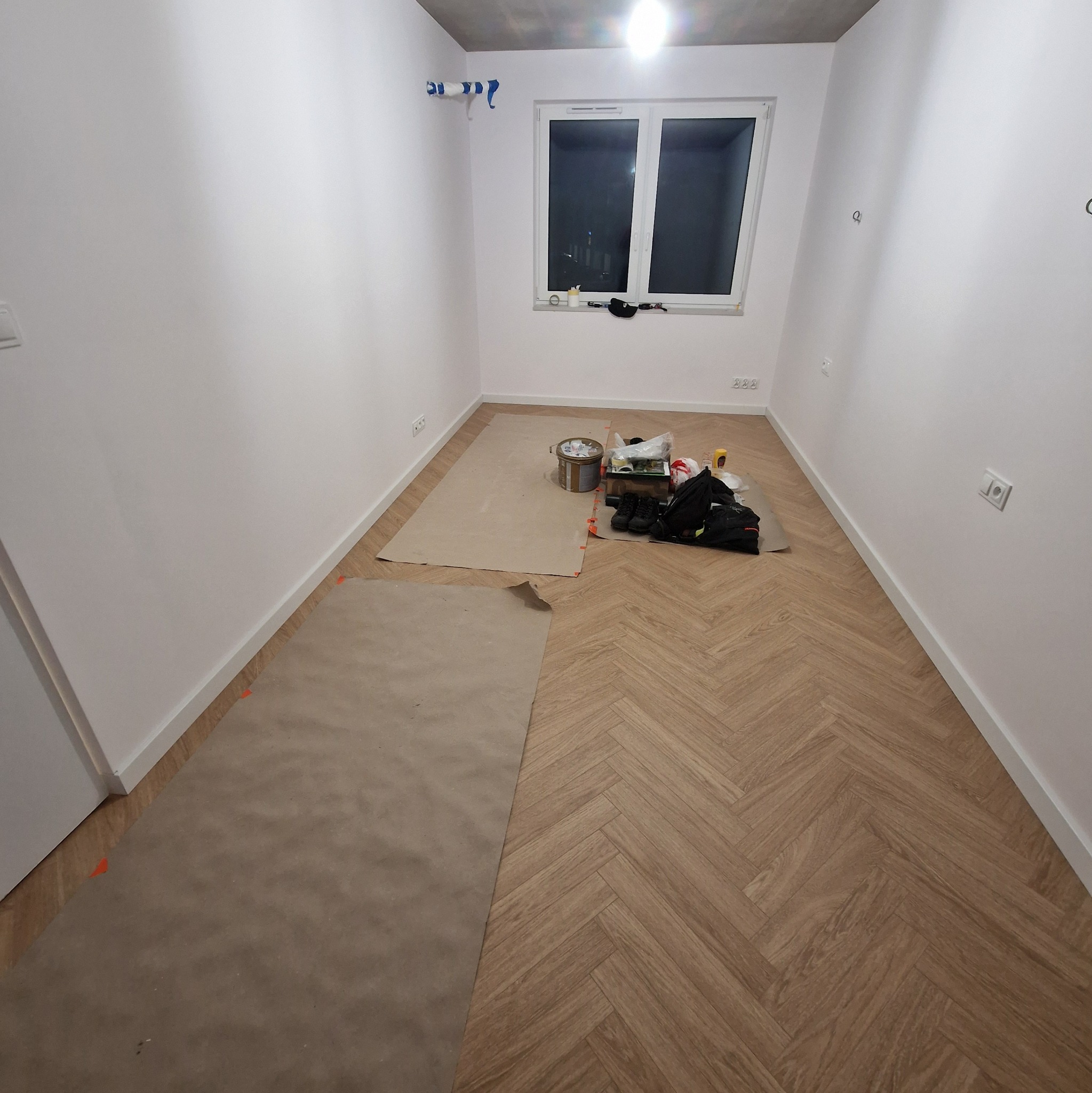 Pokój w trakcie układania parkietu w jodełkę. Część podłogi zabezpieczona brązowym papierem, widoczne narzędzia i materiały. Ściany pomalowane na biało.