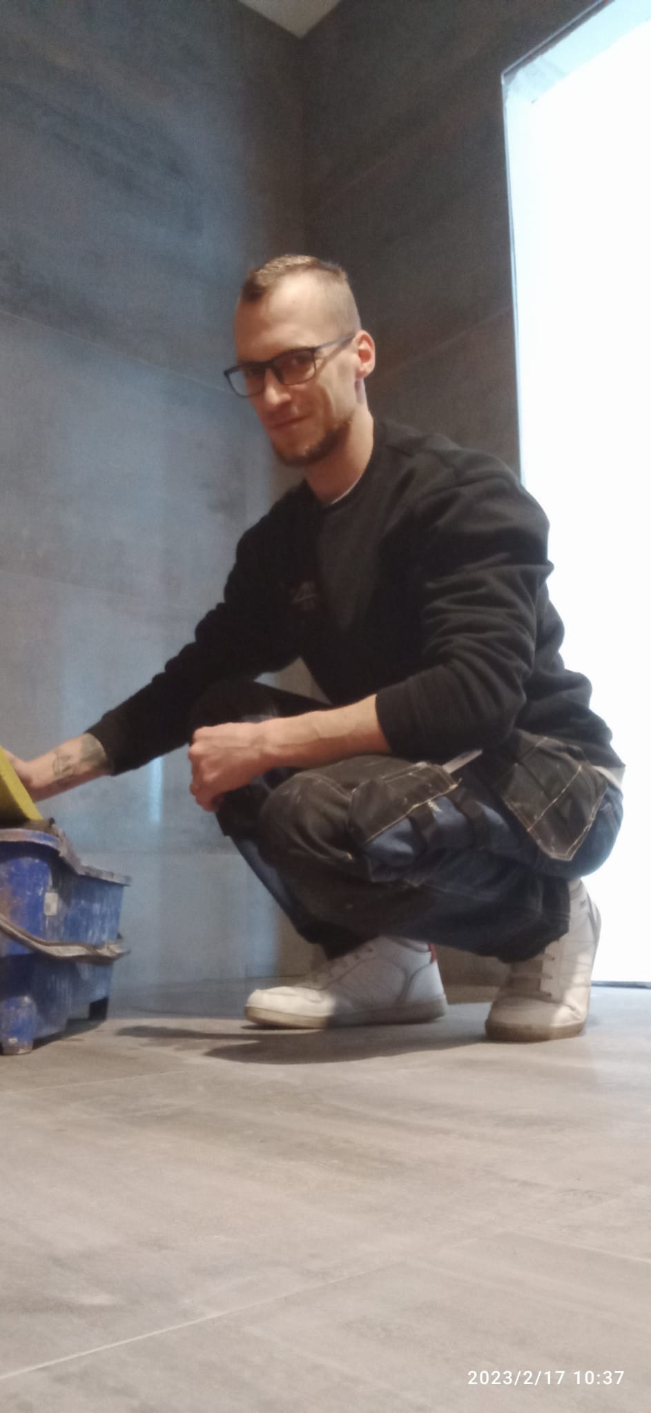 Mężczyzna w okularach, w kucki, z niebieskim wiadrem i gąbką w łazience z szarymi płytkami, prawdopodobnie podczas czyszczenia po fugowaniu.