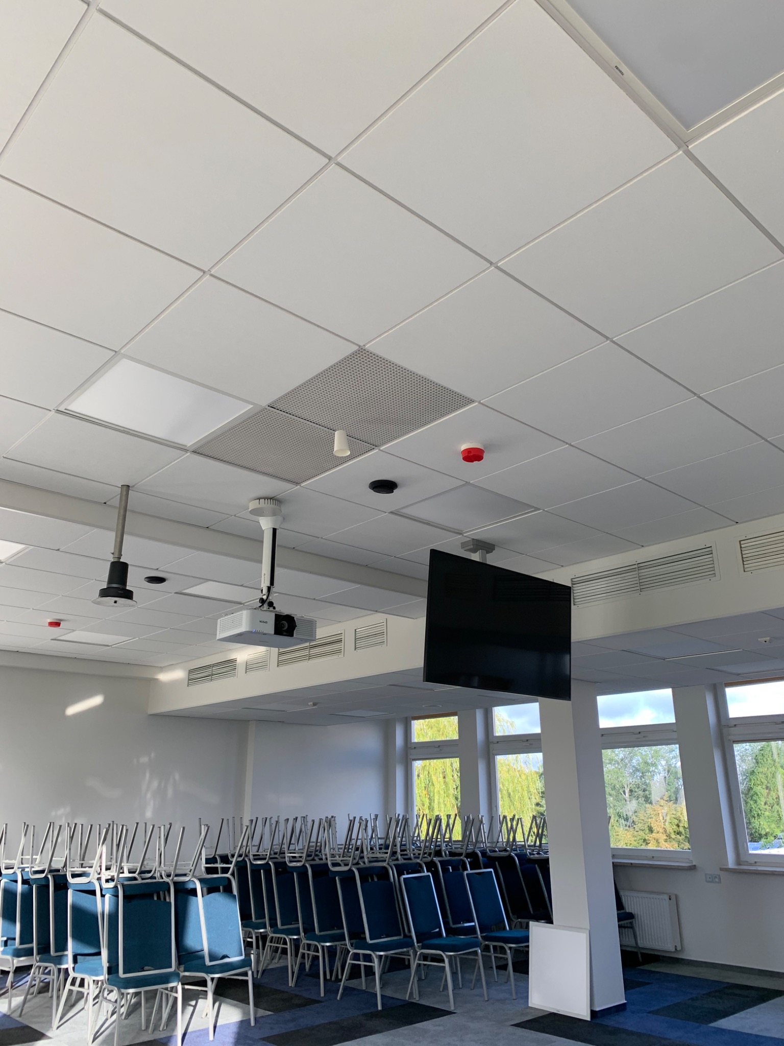 Sala konferencyjna z białym sufitem kasetonowym, projektorem, ekranem i krzesłami ustawionymi w rzędach, widok z dołu na sufit, w tle okna z widokiem na drzewa.