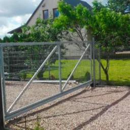 Otwarta, przesuwna brama z siatki metalowej, zamontowana na posesji z domem jednorodzinnym, widoczna skrzynka pocztowa przy słupku.