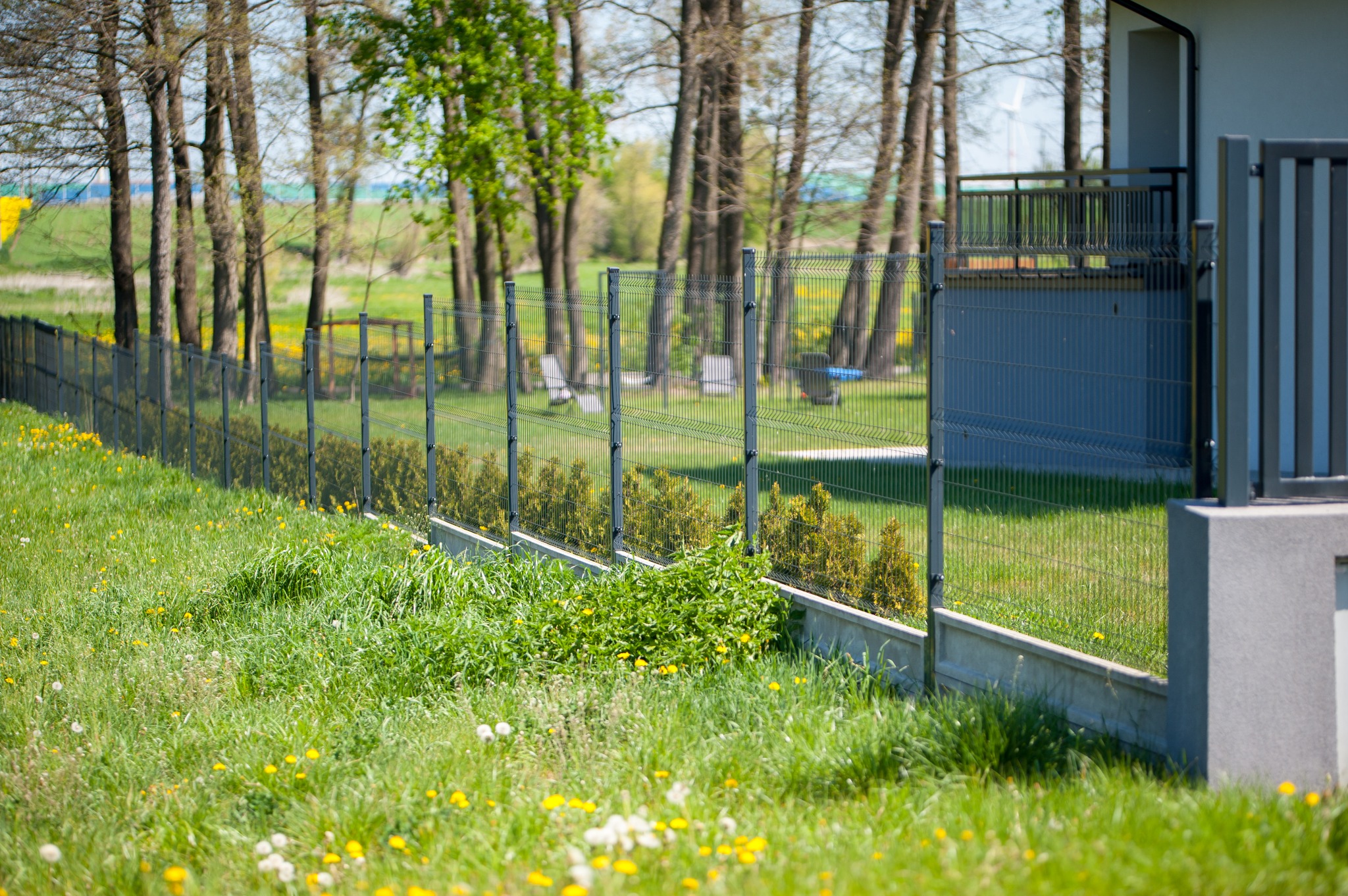 Nowoczesne, grafitowe ogrodzenie panelowe z podmurówką betonową, widok z zewnątrz na tle zielonego trawnika z żółtymi kwiatami i drzew