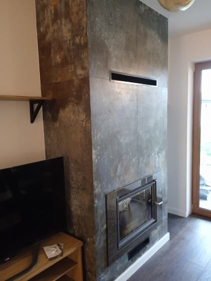 Obudowa kominka z szarych, wielkoformatowych płytek imitujących kamień, z widocznym wkładem kominkowym i poziomym, wąskim otworem wentylacyjnym, w tle telewizor i półka