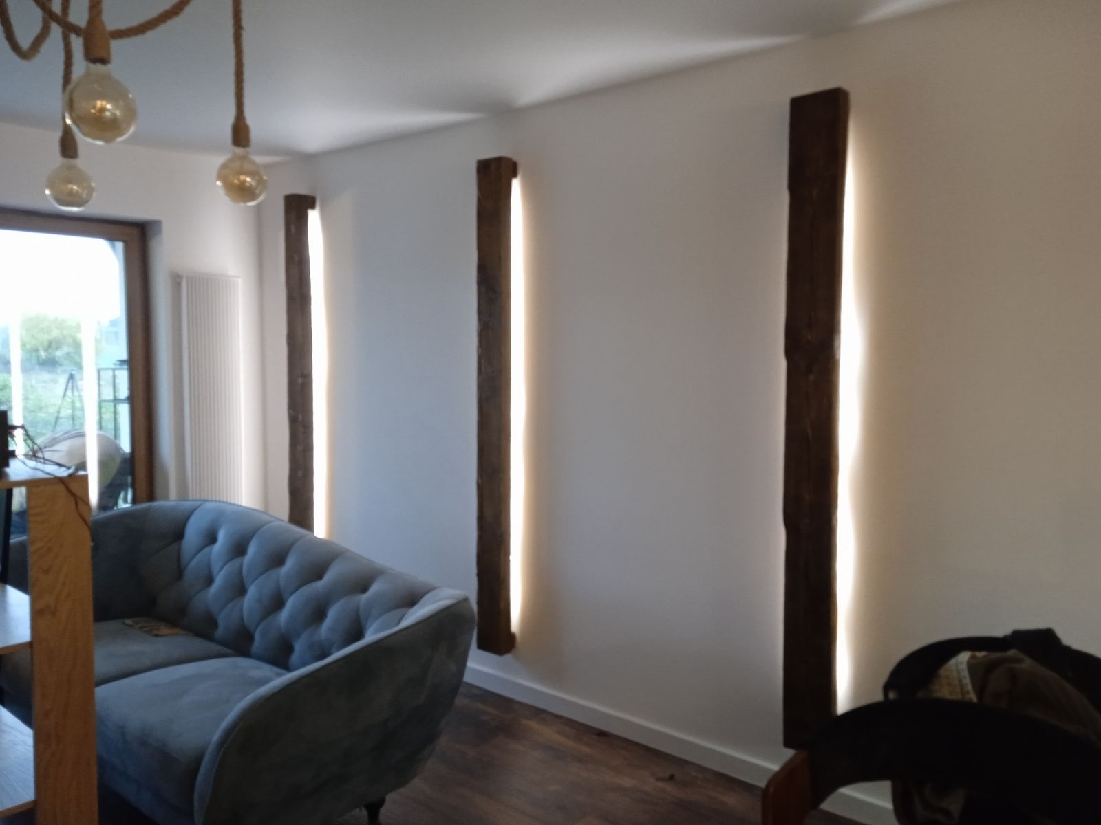 Nowoczesne oświetlenie ścienne z drewnianymi elementami i ukrytym źródłem światła, uzupełnione szarą sofą w stylu Chesterfield na drewnianej podłodze.