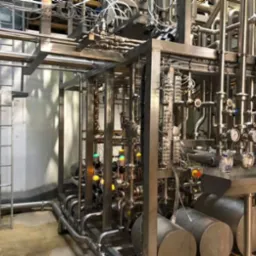 Skomplikowany system rur i zaworów ze stali nierdzewnej, z manometrami i elementami sterującymi, zamontowany w zakładzie przemysłowym, widoczna drabina z boku.