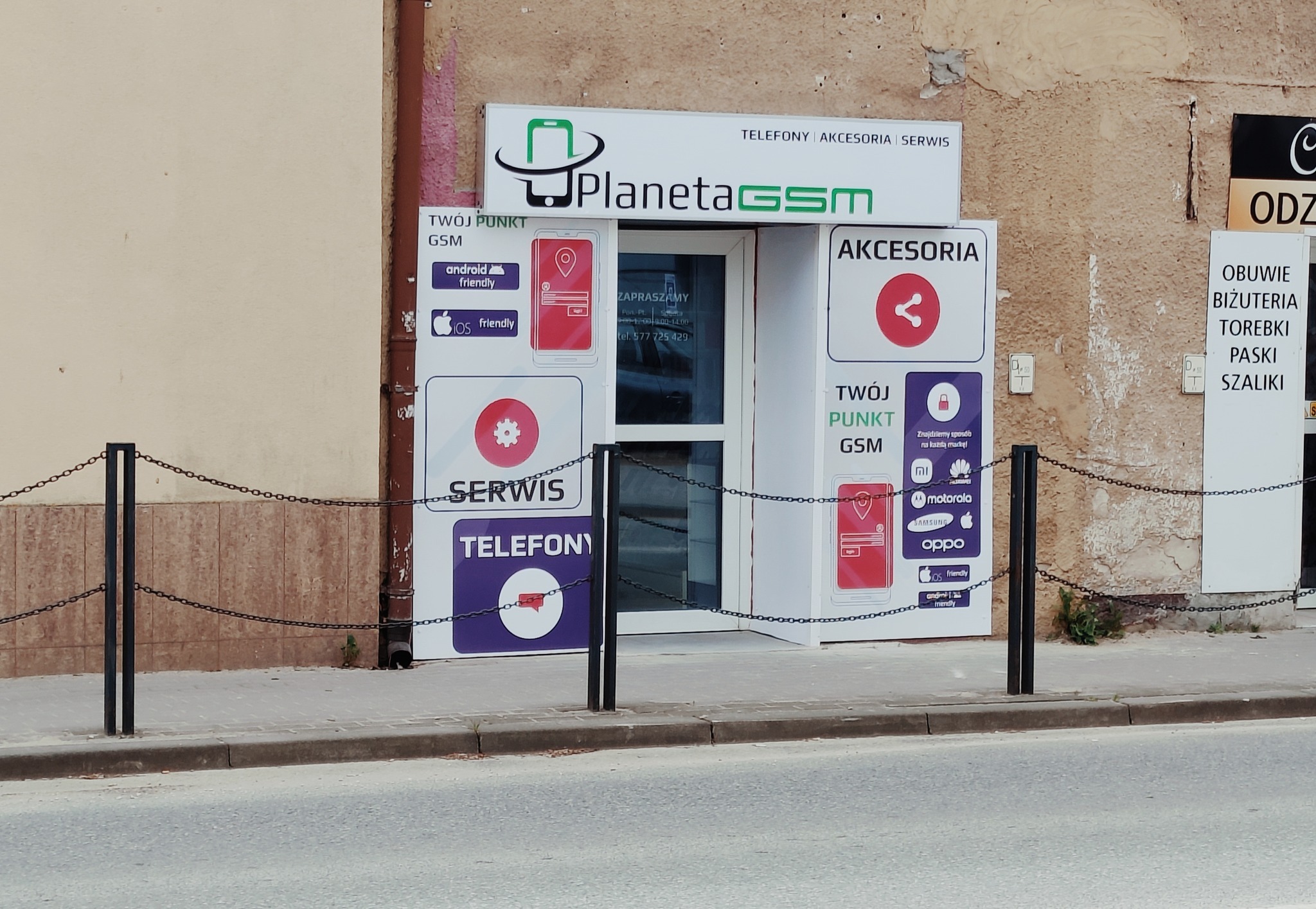 Fasada sklepu z szyldem 'Planeta GSM', oferującego akcesoria i usługi serwisowe telefonów, zabezpieczona łańcuchami. Na ścianach plakaty informacyjne o serwisie i akcesoriach, logo Android i iOS.