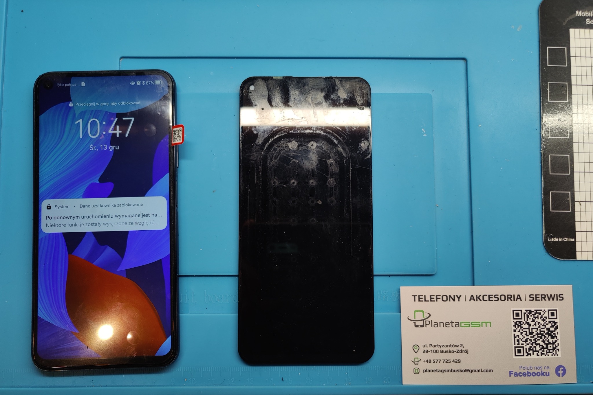 Smartfon z włączonym ekranem blokady obok zdemontowanego wyświetlacza z widocznymi śladami kleju na niebieskiej macie serwisowej, obok wizytówka serwisu.