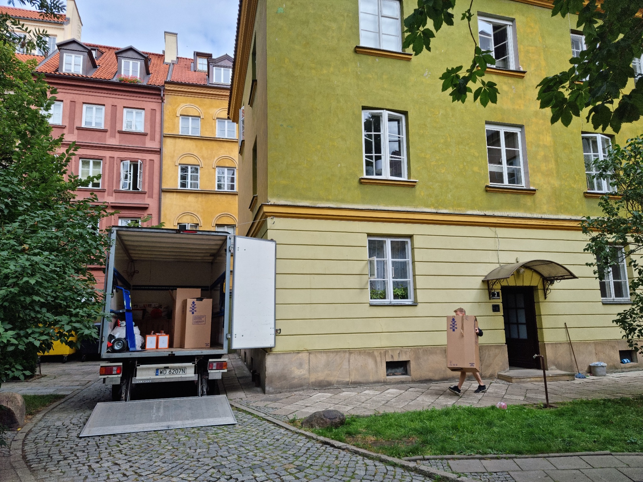 Otwarty samochód dostawczy z rampą i kartonami na tle kamienicy w Warszawie, osoba wnosząca karton do budynku.