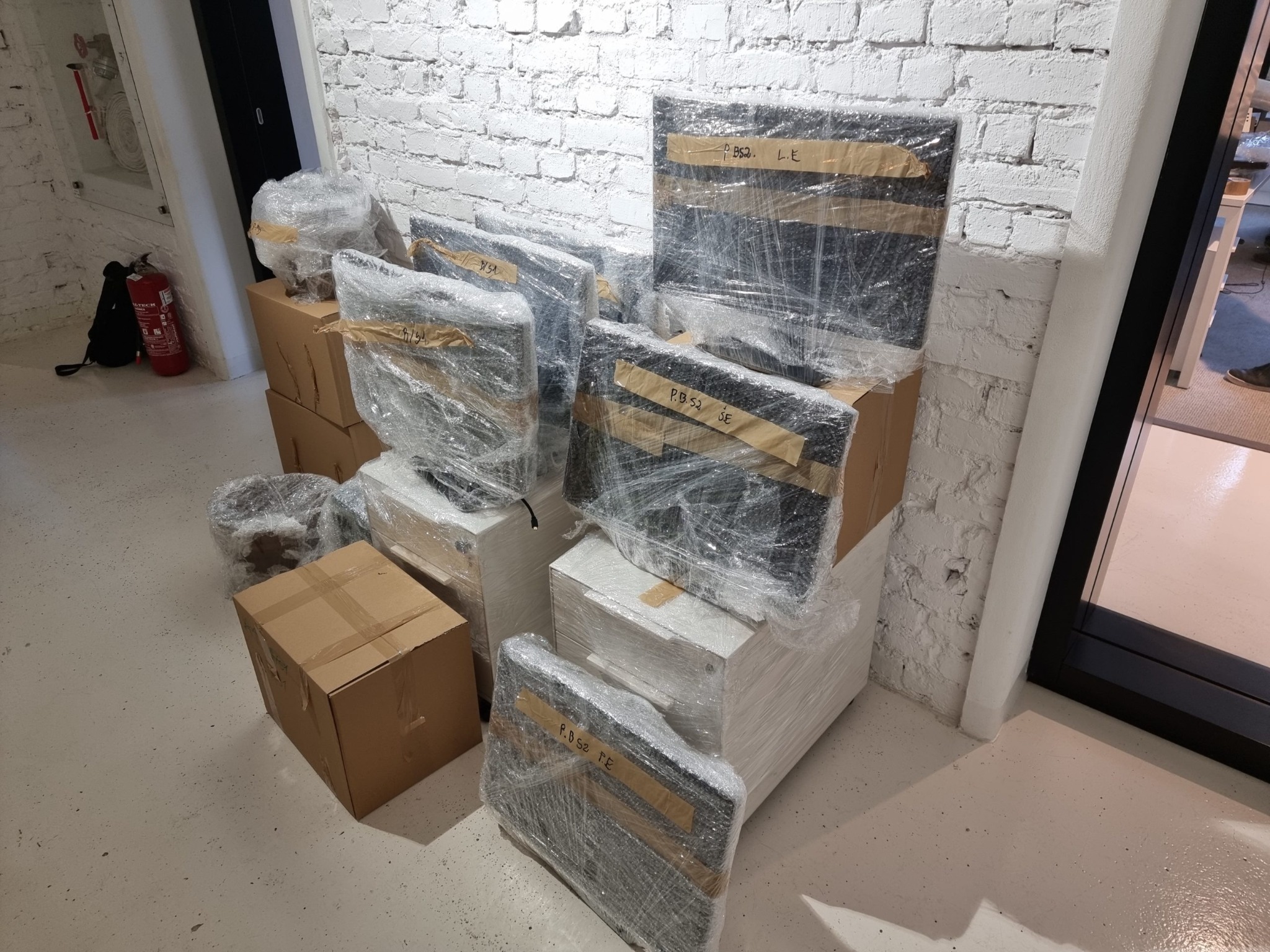 Spakowane i owinięte folią stretch monitory oraz kartony, przygotowane do transportu w pomieszczeniu z białymi ścianami z cegły i jasną podłogą.
