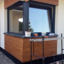 Wykończona wnęka okienna z imitacją drewna na elewacji budynku, widoczne rusztowanie malarskie, puszki z farbą i narzędzia na parapecie.