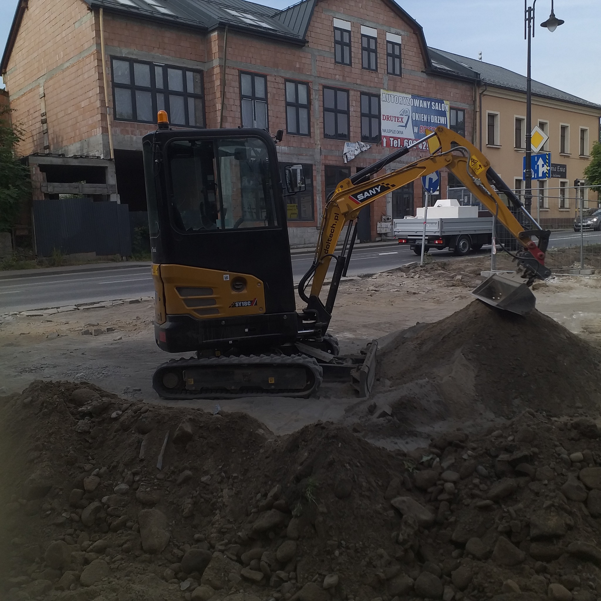 Żółto-czarna mini koparka SANY SY16C z łyżką na hałdzie ziemi i kamieni, w tle budynek w trakcie budowy i ruch uliczny.