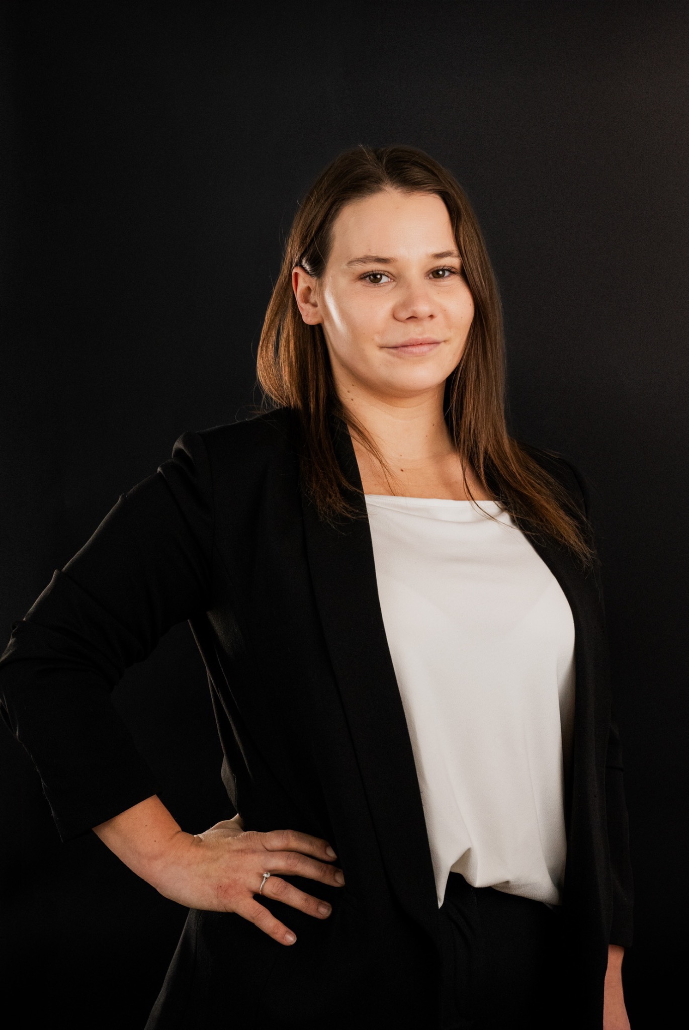 Portret kobiety w czarnym żakiecie i białej bluzce na ciemnym tle, z ręką na biodrze i pierścionkiem na palcu serdecznym.