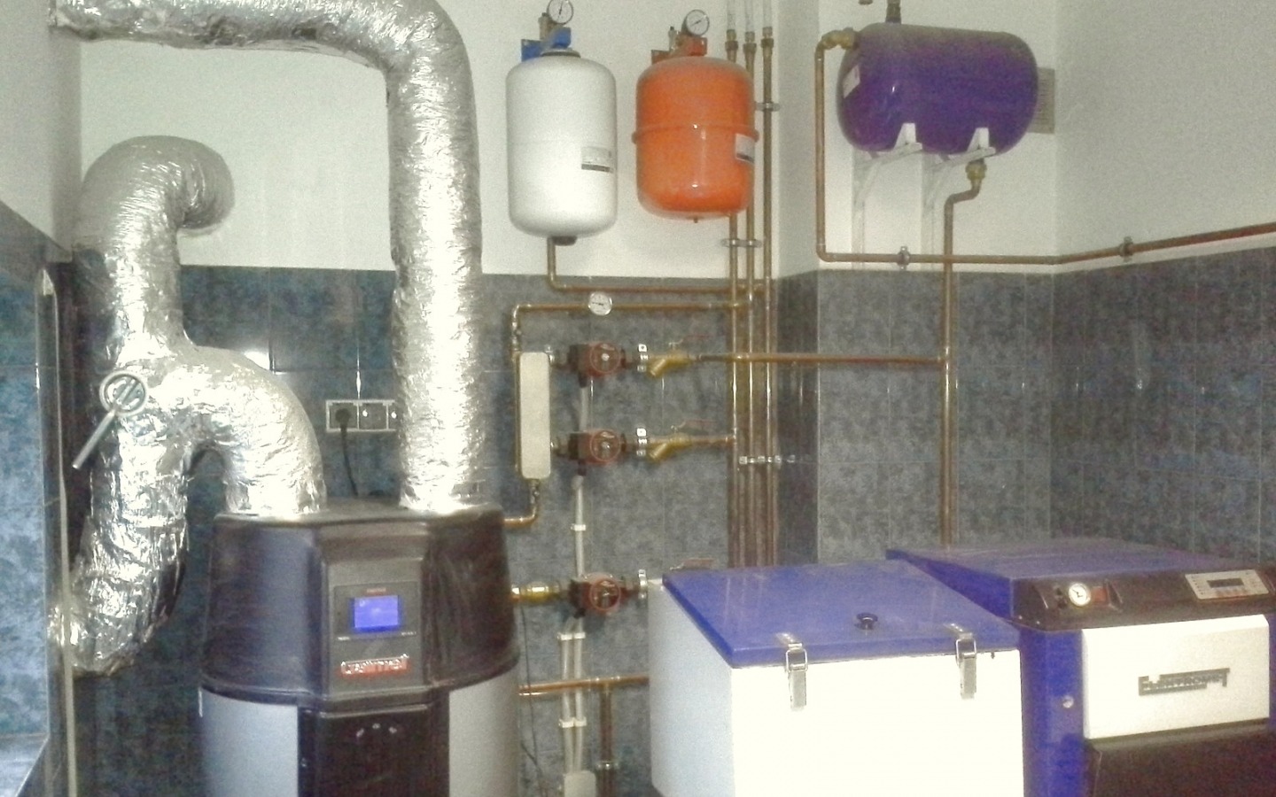 Instalacja grzewcza z kotłem, rurami miedzianymi i zbiornikami wyrównawczymi w pomieszczeniu z wykończeniem ceramicznym.