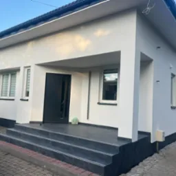 Wykończona elewacja domu jednorodzinnego z białym tynkiem strukturalnym, czarną stolarką okienną i drzwiami wejściowymi, oraz schodami wyłożonymi ciemnoszarymi płytkami imitującymi kamień.